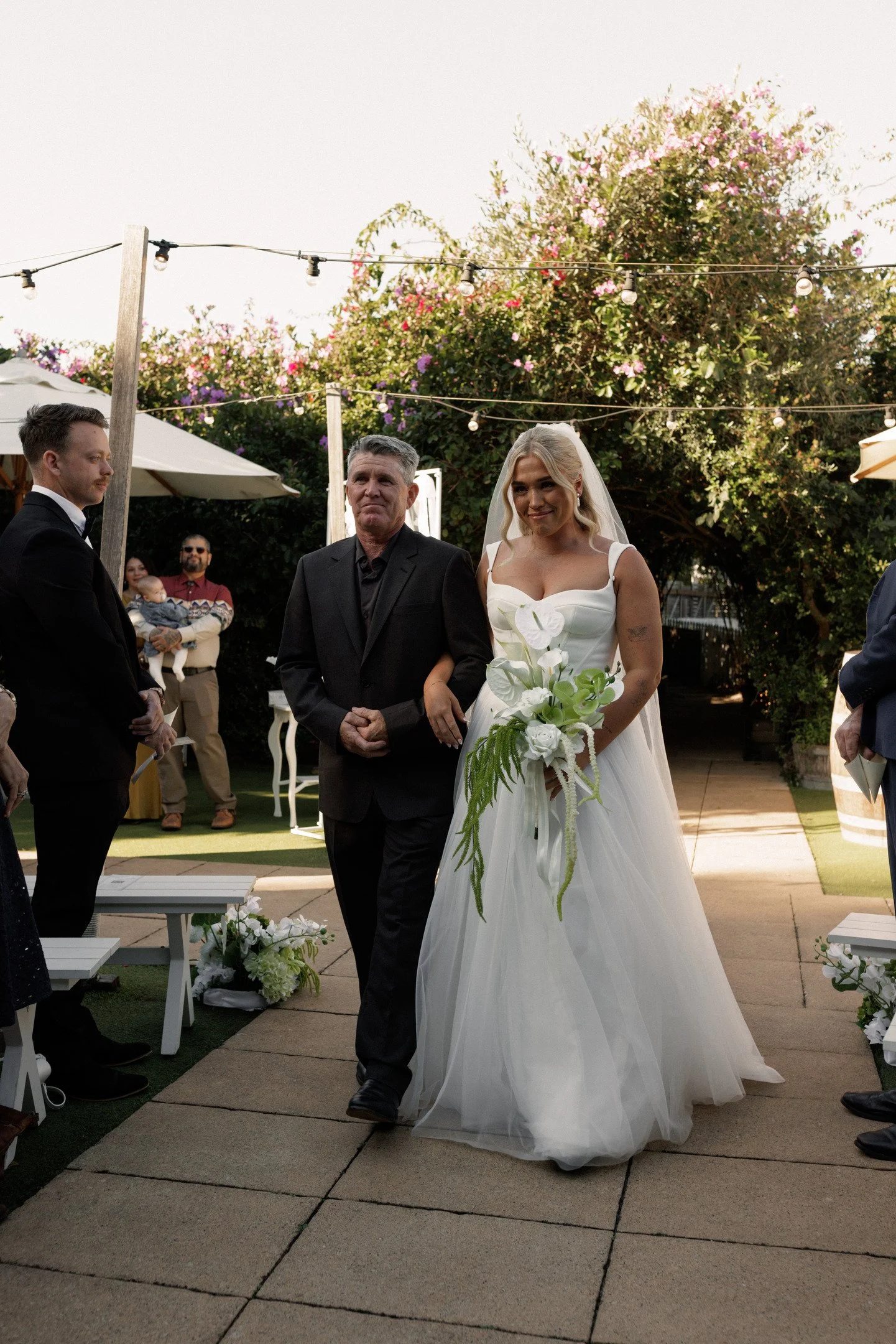 A bride in a white wedding gown holding a bouquet of white flowers, walking down an outdoor aisle with a man in a black suit, while guests look on during a wedding ceremony in the yard with string lights, trees, and flowers.