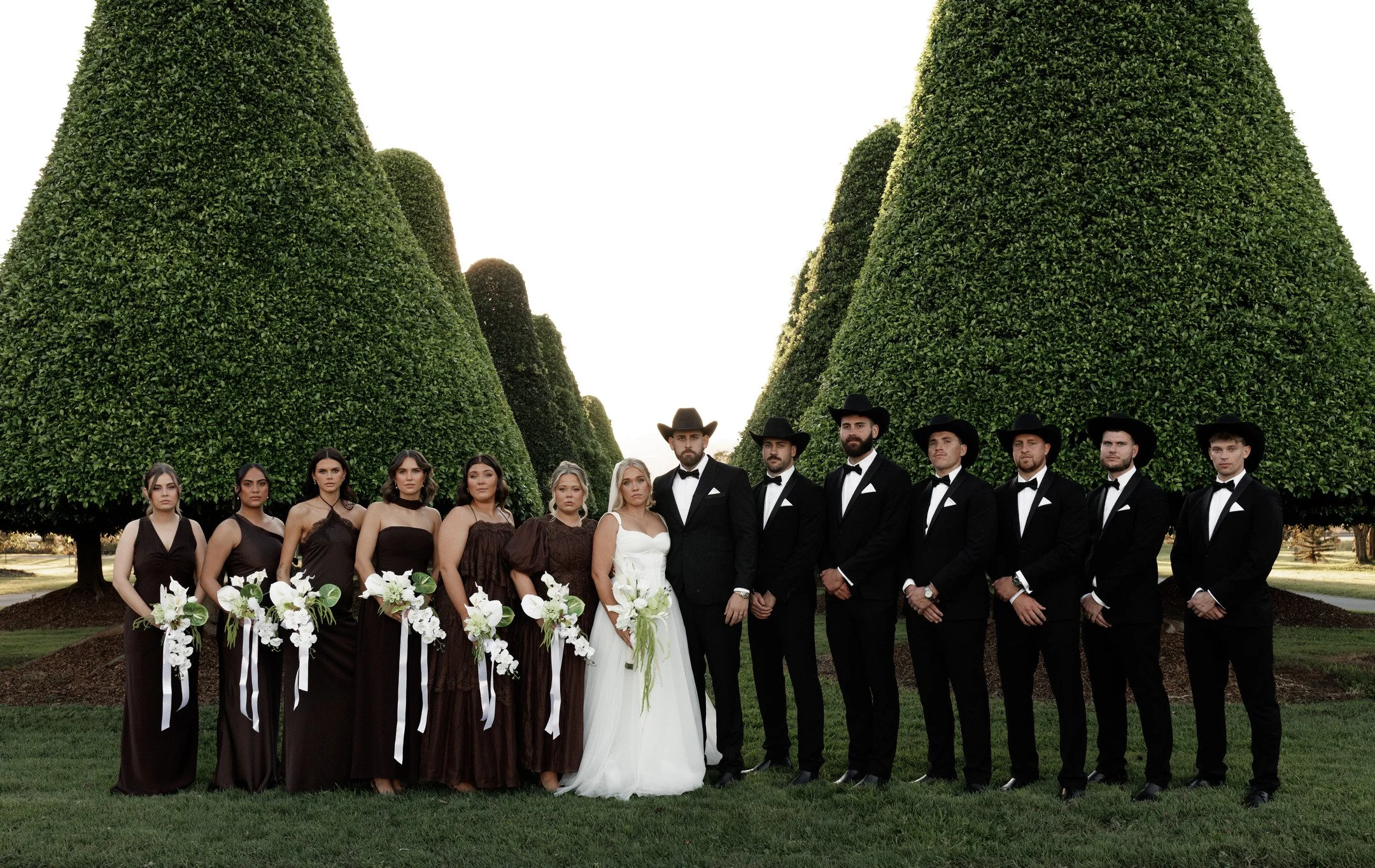 A large group of people dressed in formal attire, standing outdoors in front of tall, manicured green trees, likely at a wedding or formal event.