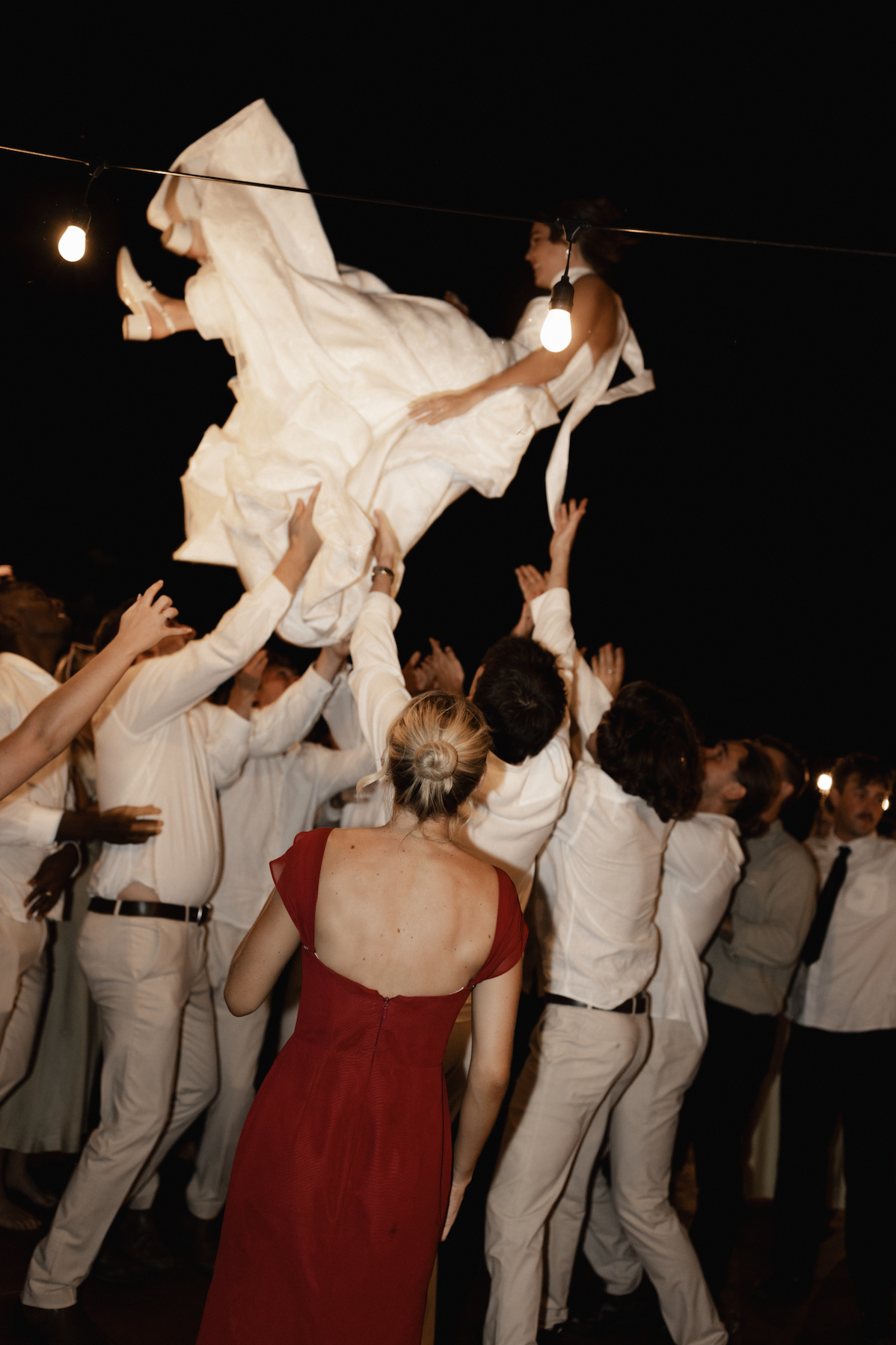 A group of people lifting a woman in a white dress during a celebration or party at night, with string lights overhead.