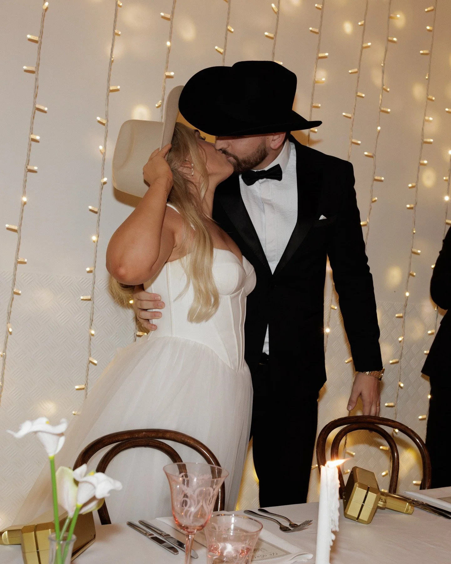 A bride and groom sharing a kiss at their wedding reception, with string lights in the background. The bride is wearing a white wedding dress and a white hat, while the groom is dressed in a black tuxedo with a bow tie and a large black hat.