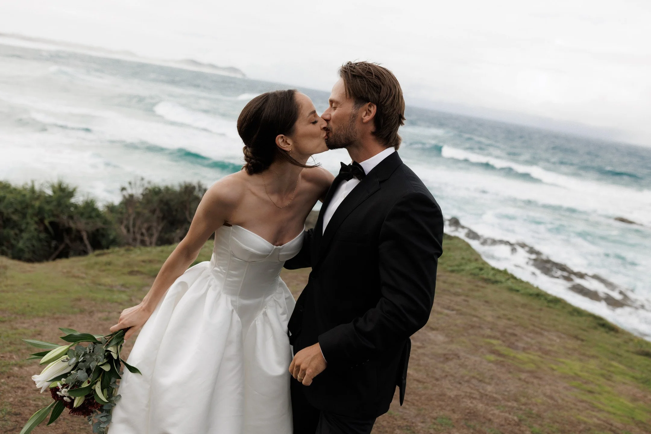 A bride and groom are kissing outdoors by the ocean. The bride is holding a bouquet of white lilies and greenery, and she is wearing a white strapless wedding dress. The groom is dressed in a black tuxedo with a bow tie. The background features waves and a cloudy sky.