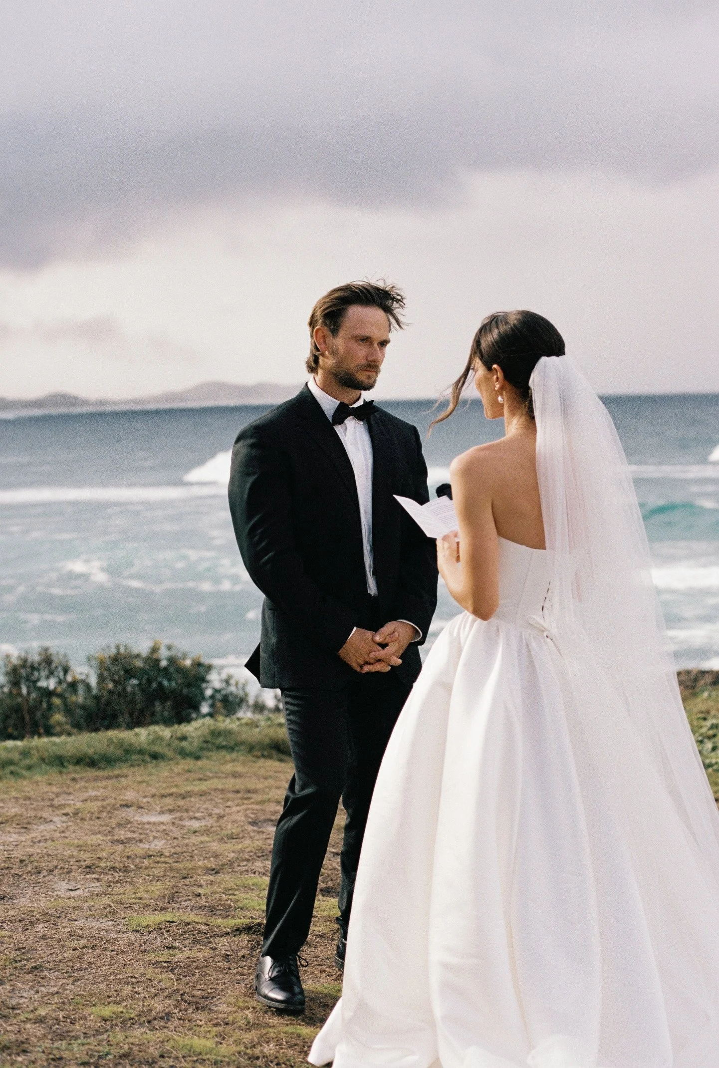 A bride and groom stand facing each other by the ocean, with the bride holding a paper and wearing a white dress and veil, while the groom wears a black tuxedo with a bow tie, under a cloudy sky.