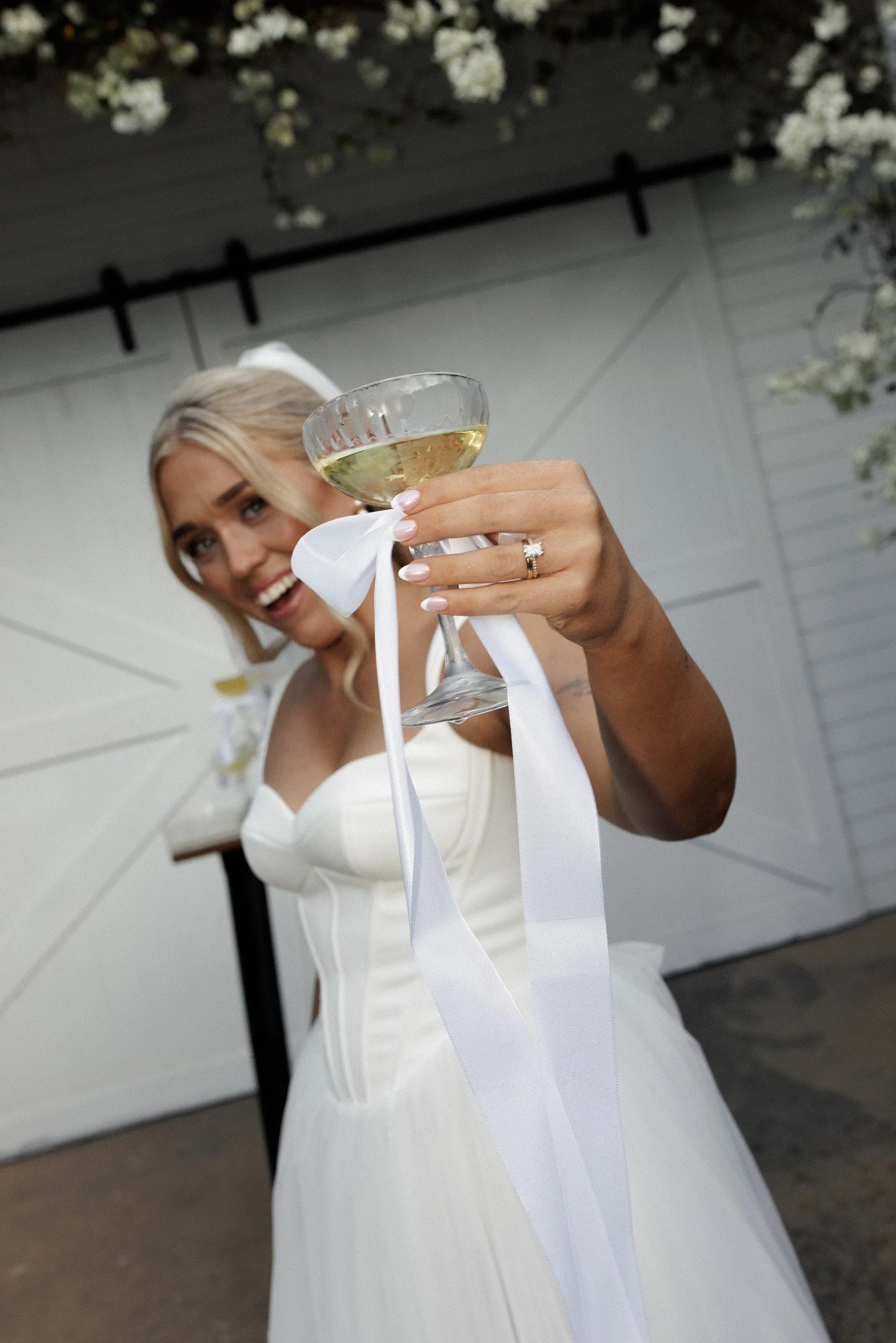 A blonde woman in a white dress is smiling and holding up a glass of white wine or champagne at a celebration or wedding event.