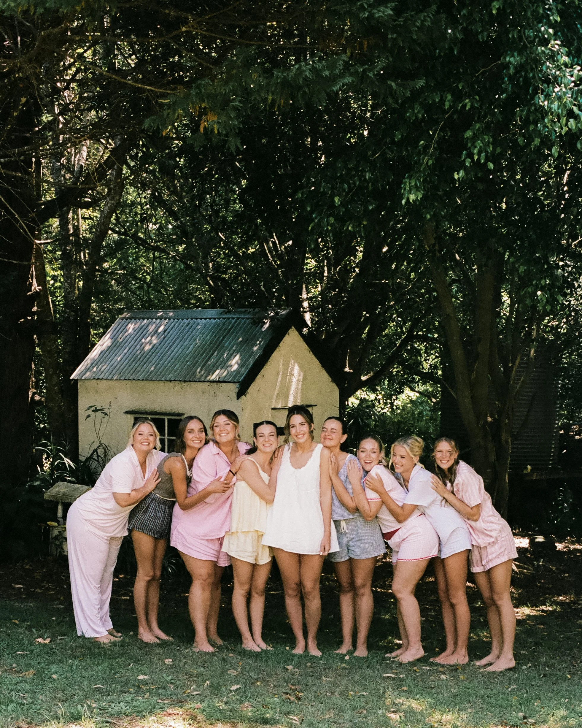 Group of nine women standing outdoors in pajamas, smiling, with trees and a small white building with a metal roof in the background.