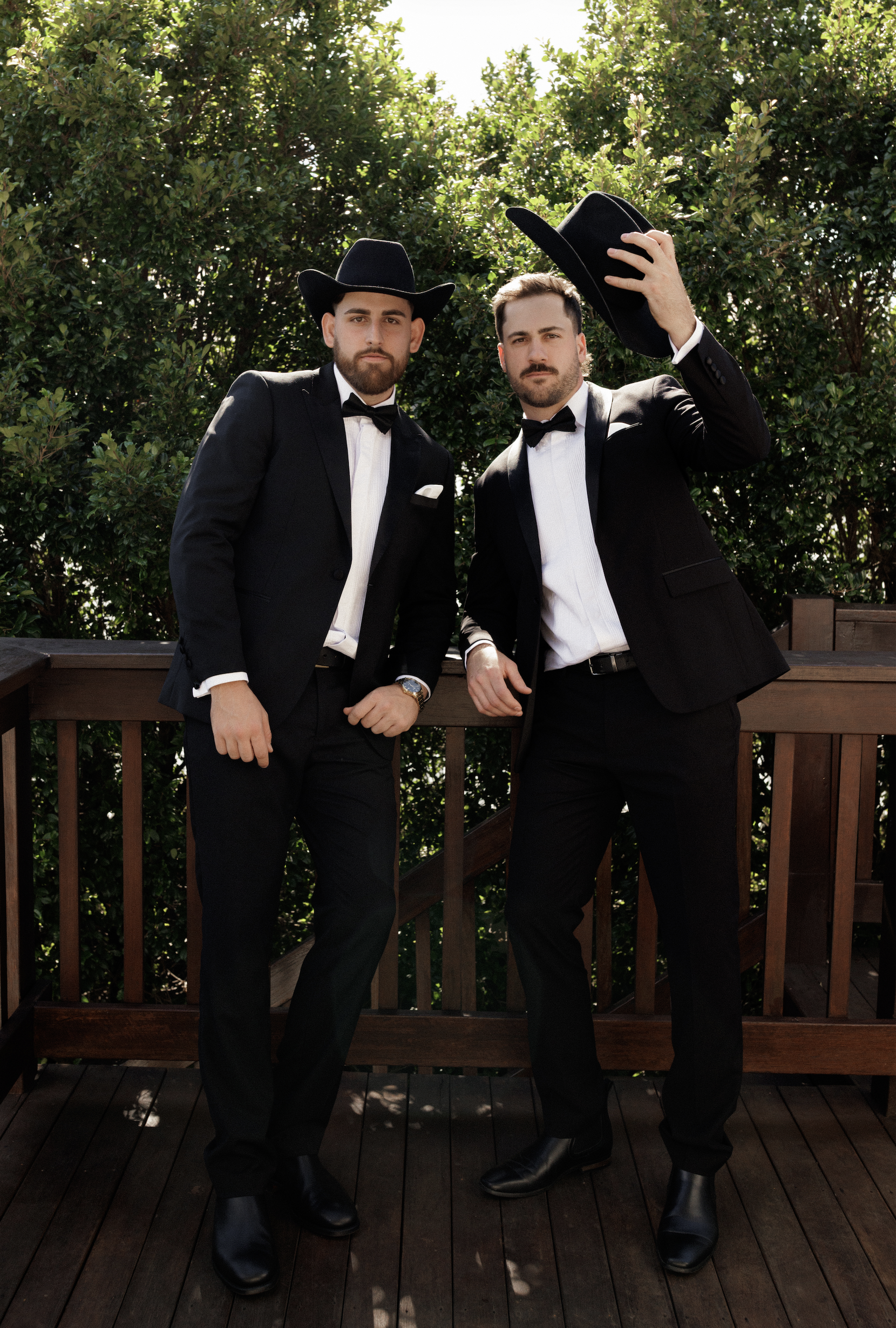 Two men dressed in tuxedos and cowboy hats standing on a wooden deck in front of green bushes, one with the hat held in hand.