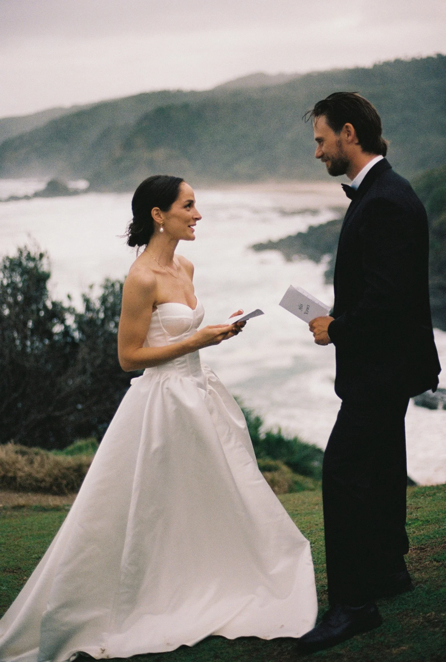A bride and groom standing outdoors near the coast, holding papers and looking at each other, with mountains and the ocean in the background.