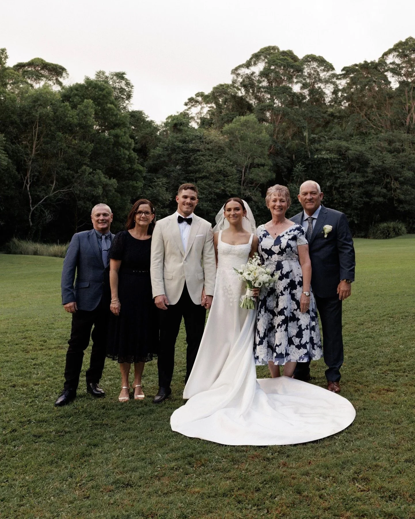 A group of six people, including a bride and groom, standing on a grassy field with trees in the background during the daytime. The bride is wearing a white wedding gown and holding a bouquet, while the groom is dressed in a white tuxedo with a black bow tie. The other four people, appearing as family members or friends, are dressed in formal attire and are smiling for the photo.