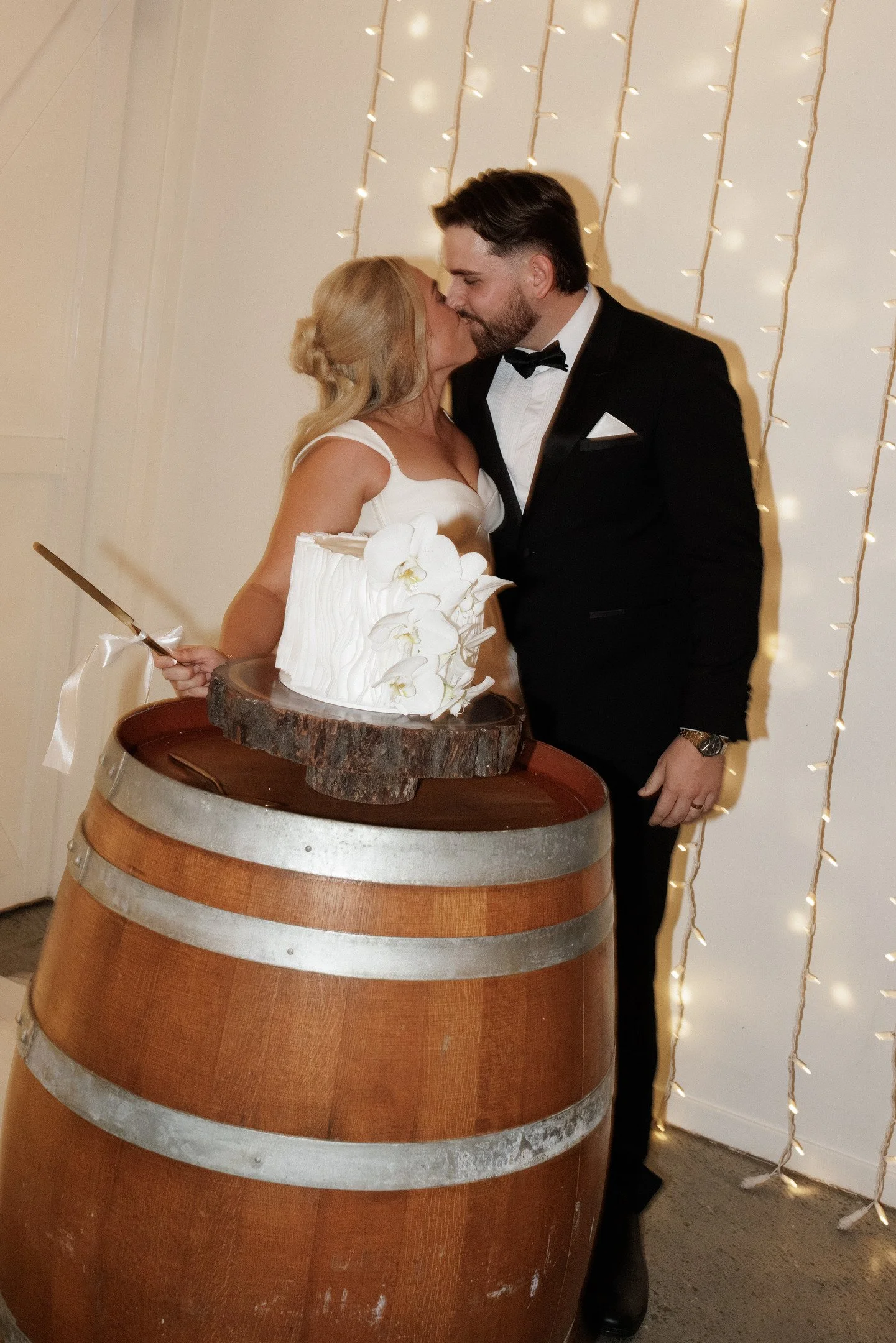 A bride and groom kissing at their wedding reception. The bride is holding a knife and standing next to a wedding cake on a barrel. The background features string lights.