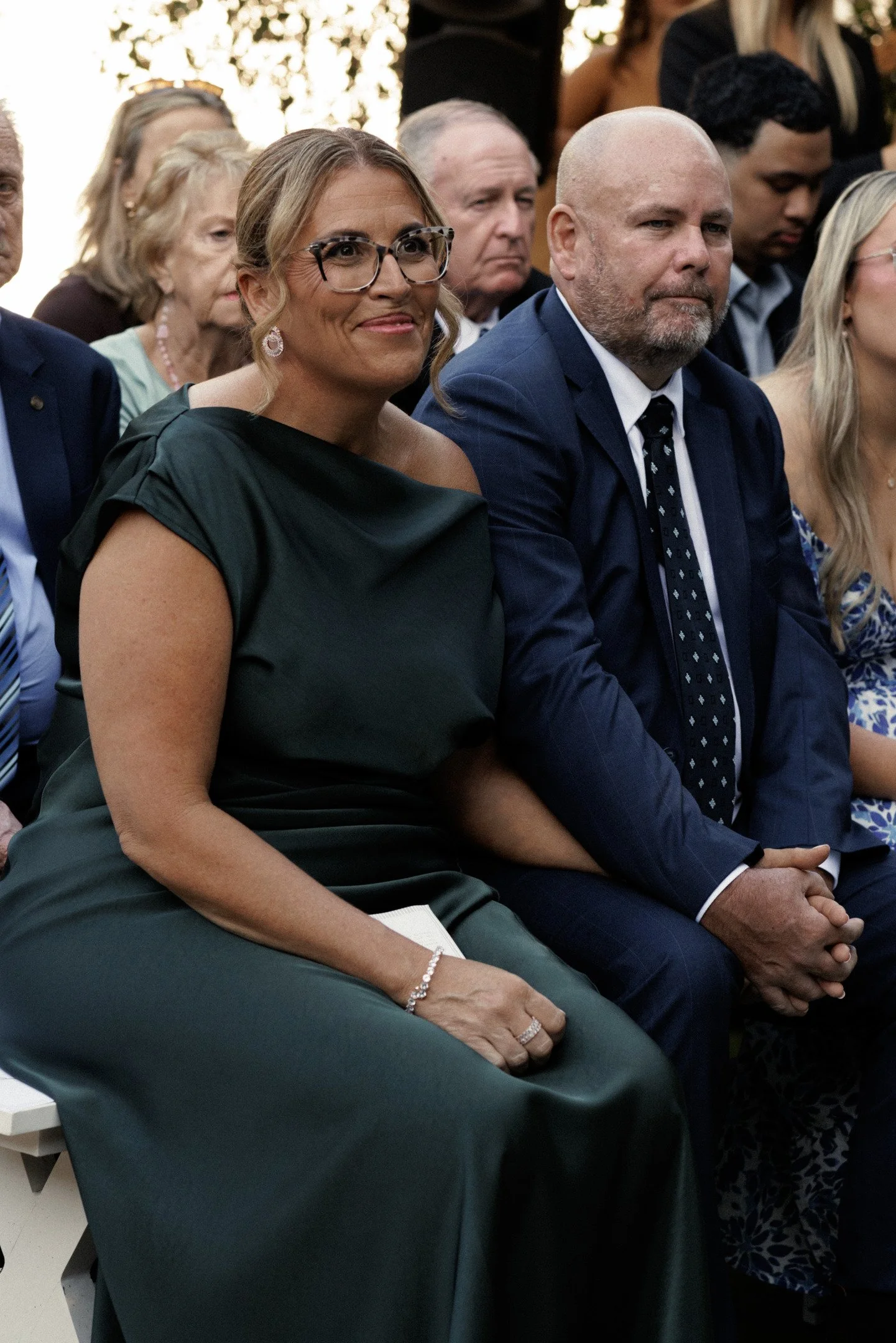 Group of adults seated at an outdoor event, dressed in formal attire, with people of various ages in the background. The focus is on a woman wearing glasses and a dark green dress, and a man in a blue suit with a patterned tie, both looking forward.