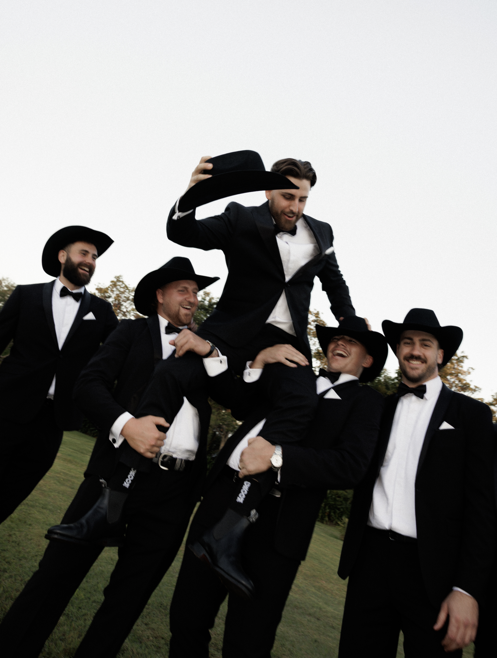 Group of men in tuxedos and cowboy hats celebrating outdoors, one man is being lifted up, smiling and holding a cowboy hat.