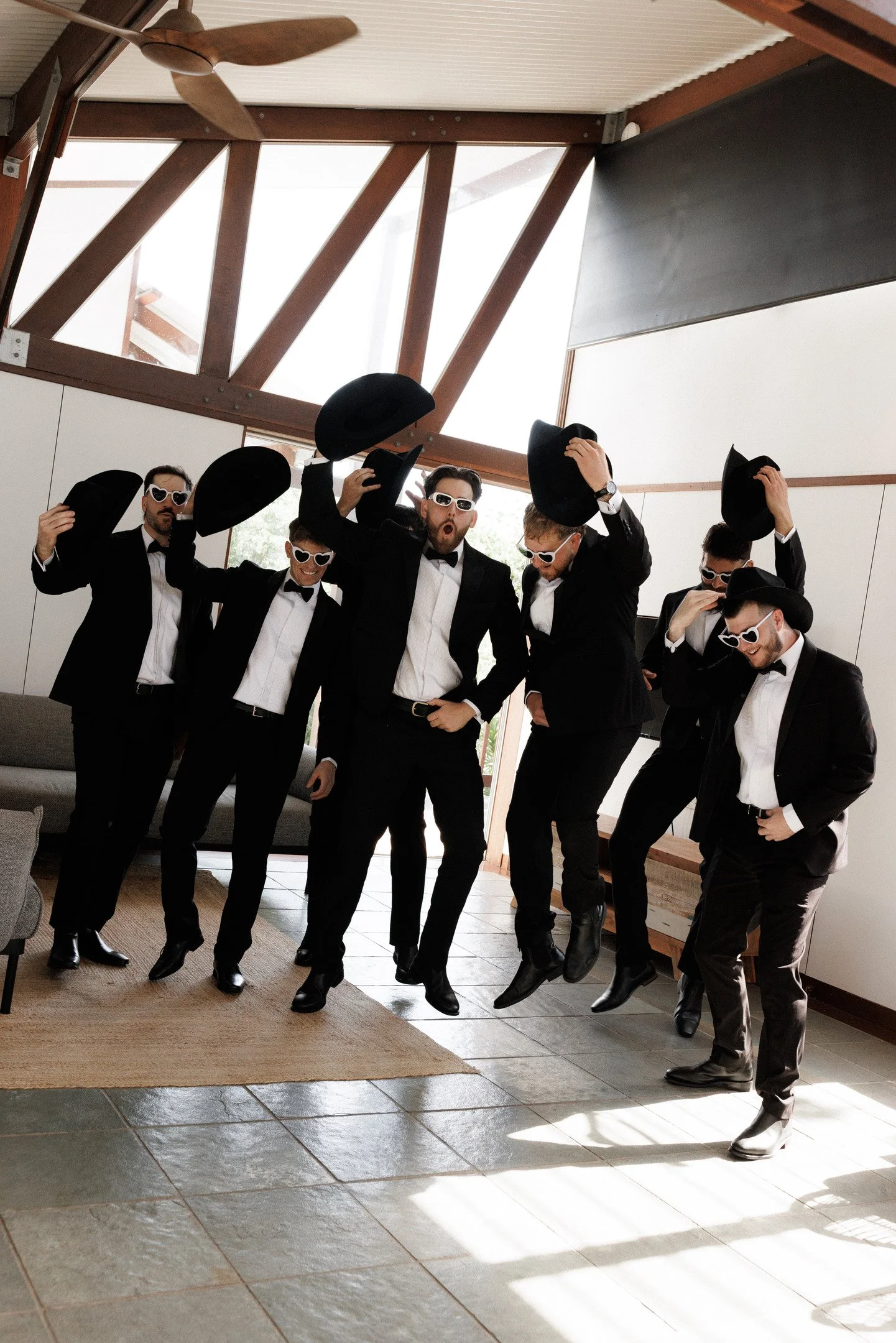 A group of six men dressed in black tuxedos and white shirts are celebrating indoors. They are wearing sunglasses and holding up black hats, with some tipping their hats or adjusting them, in a playful manner. The room has a high ceiling with wooden 