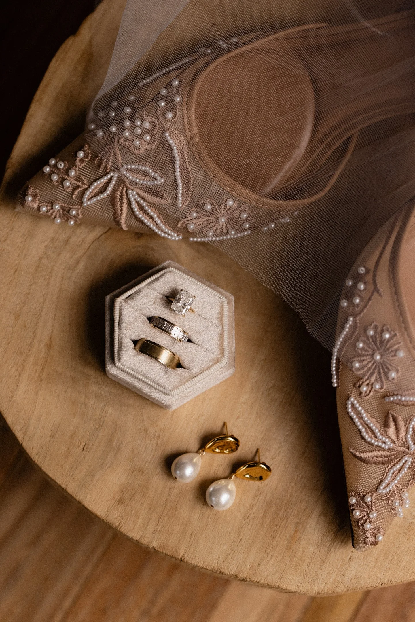 A round wooden table holding jewelry including pearl earrings, rings, and a jewelry box, partially covered with a silky pink fabric embroidered with pearls and floral designs.