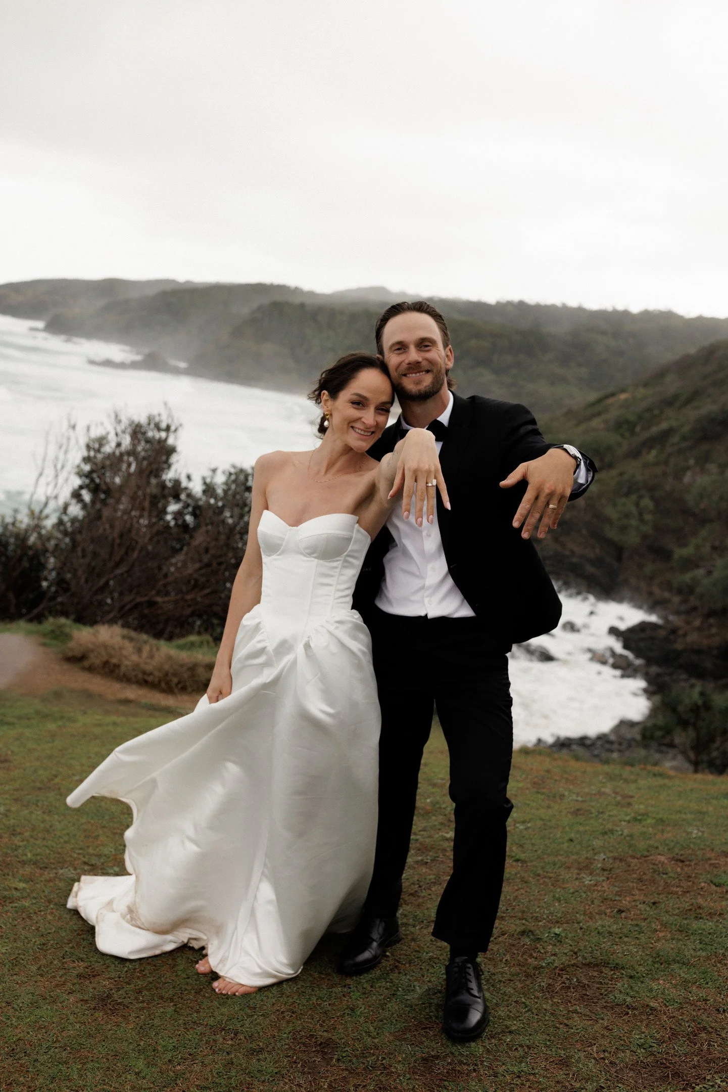 A happy couple in wedding attire standing outdoors by the ocean with misty hills in the background, smiling and showing off wedding rings.