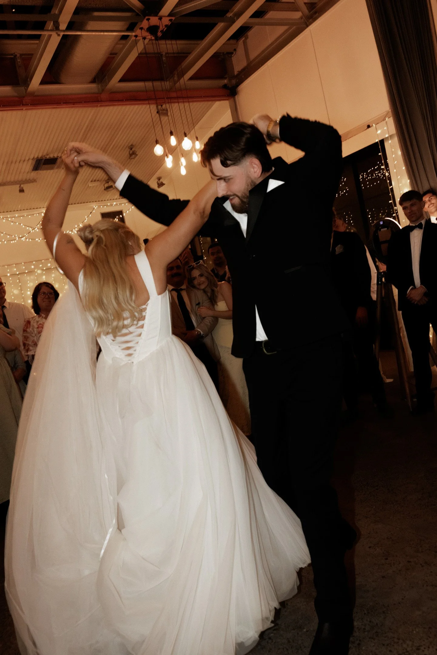 A couple dancing at a formal event, with the woman wearing a white wedding gown and the man in a black tuxedo, surrounded by onlookers and string lights in the background.