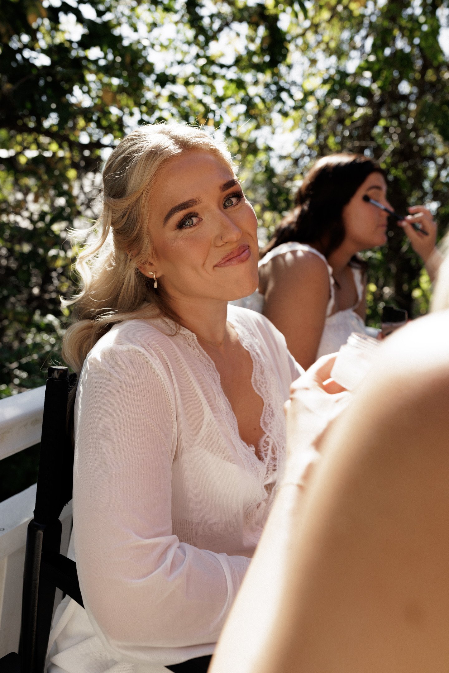 A blonde woman smiling at the camera while sitting outdoors with two women in the background, one applying makeup, amidst green foliage.