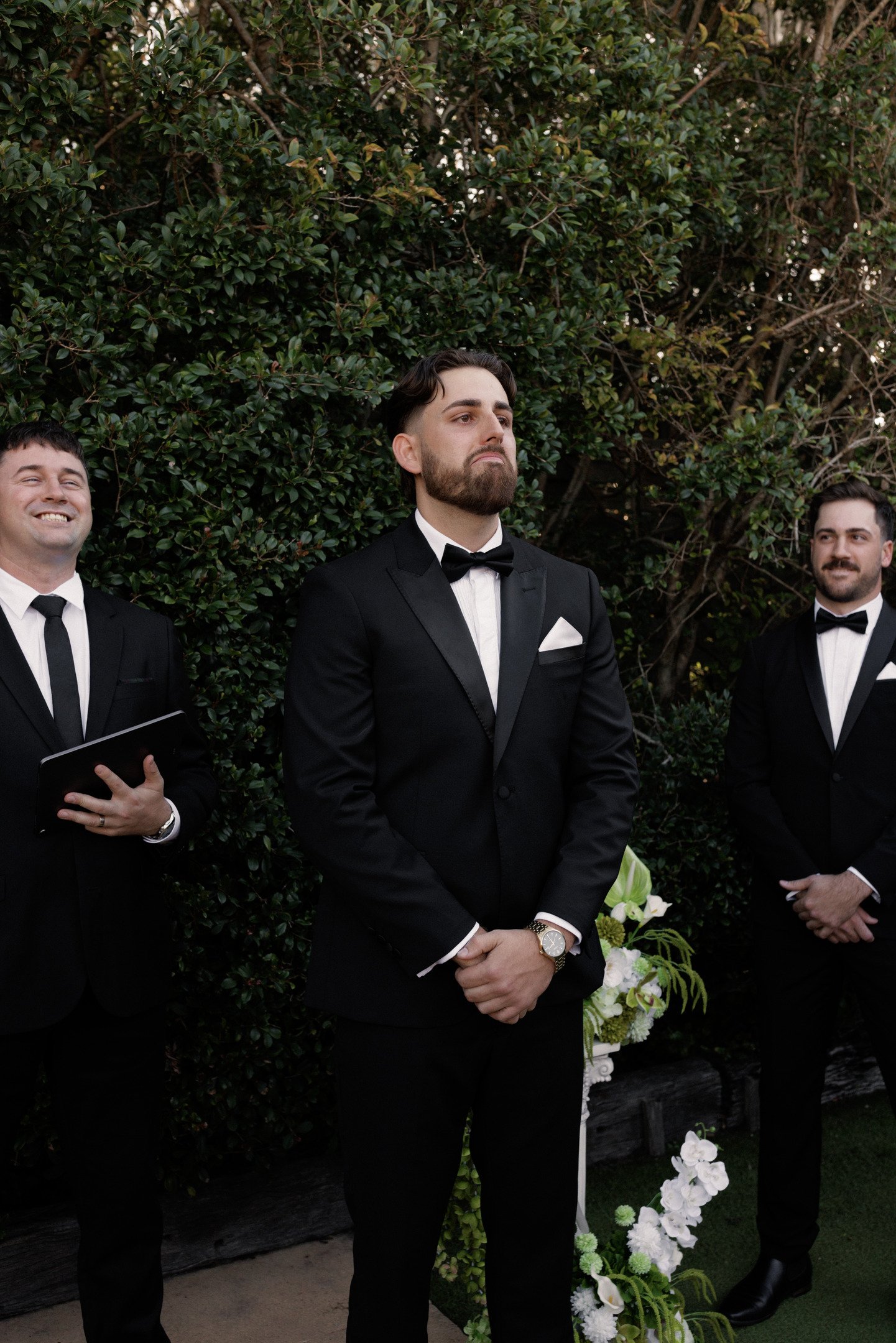 Three men dressed in black tuxedos with bow ties standing outdoors in front of a leafy green hedge. The man in the center has dark hair, a beard, and is looking serious, while the other two men are smiling.