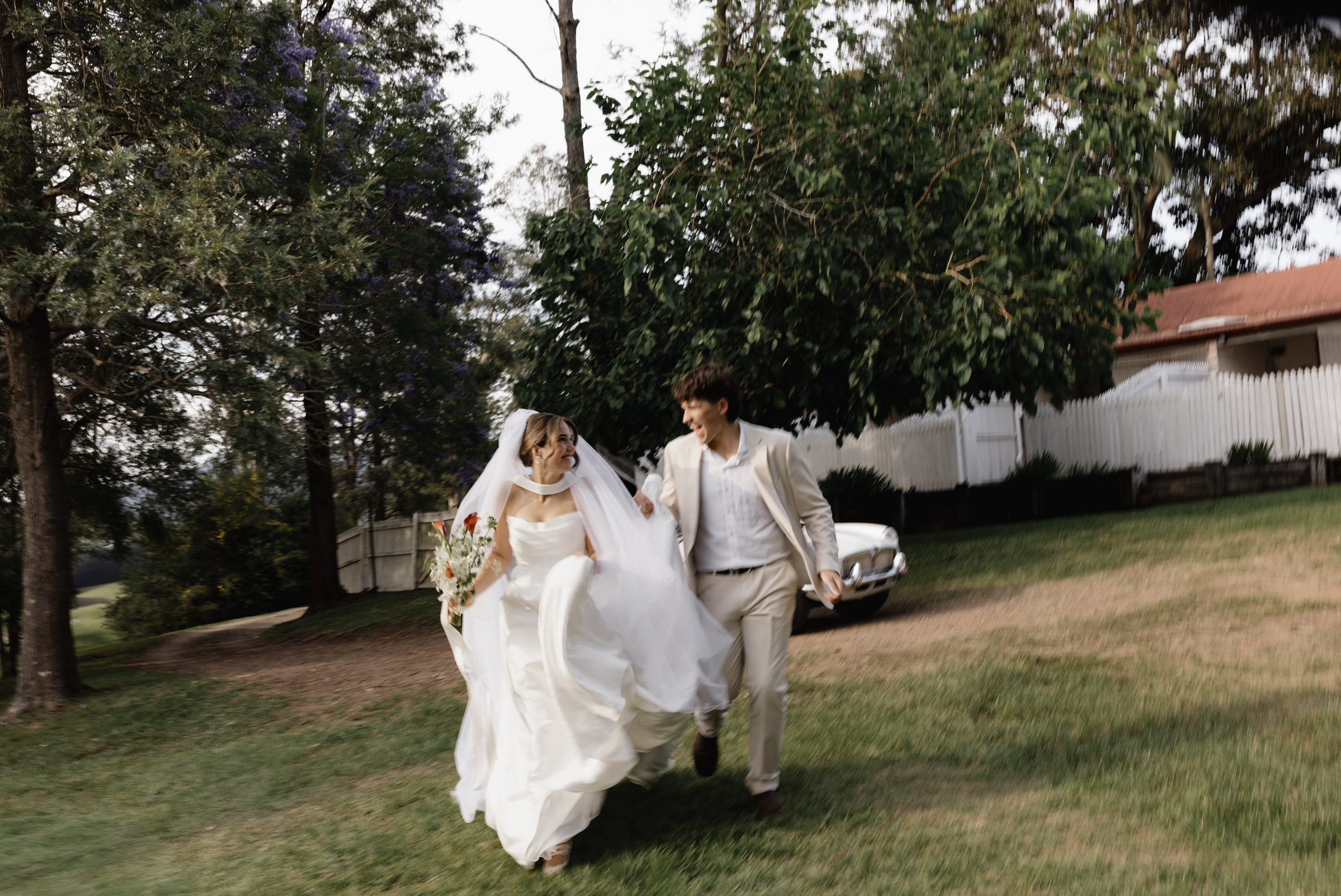 Bride and groom running outdoors on grass, smiling and laughing, with wedding attire; trees, white fence, and house in background.