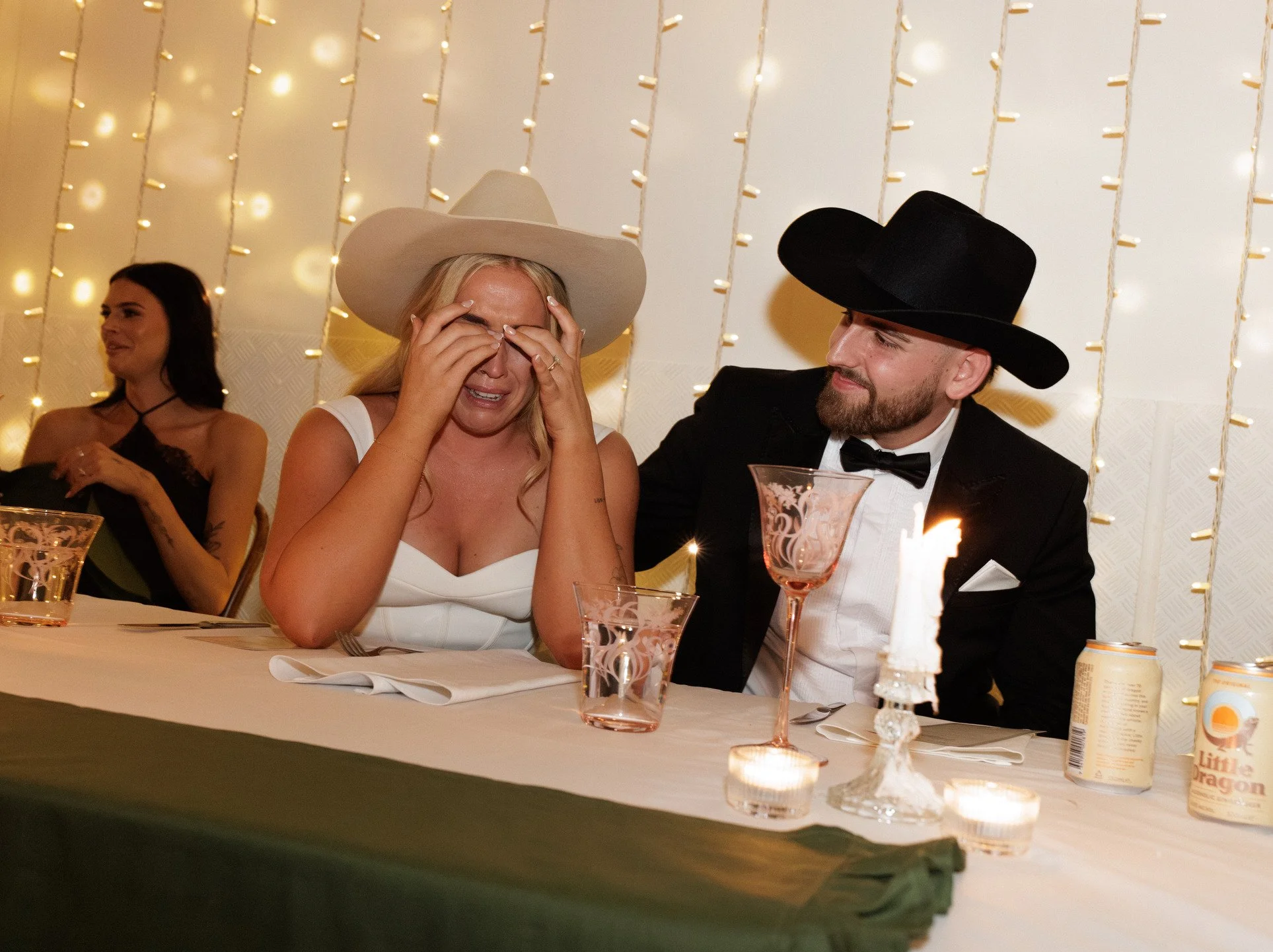 A woman wearing a white dress and a wide-brimmed hat sits at a table, visibly emotional or tearful, with a man in a suit and fedora hat smiling nearby. The table is decorated with pink glasses, candlelight, and food and drink items, and there is a ba