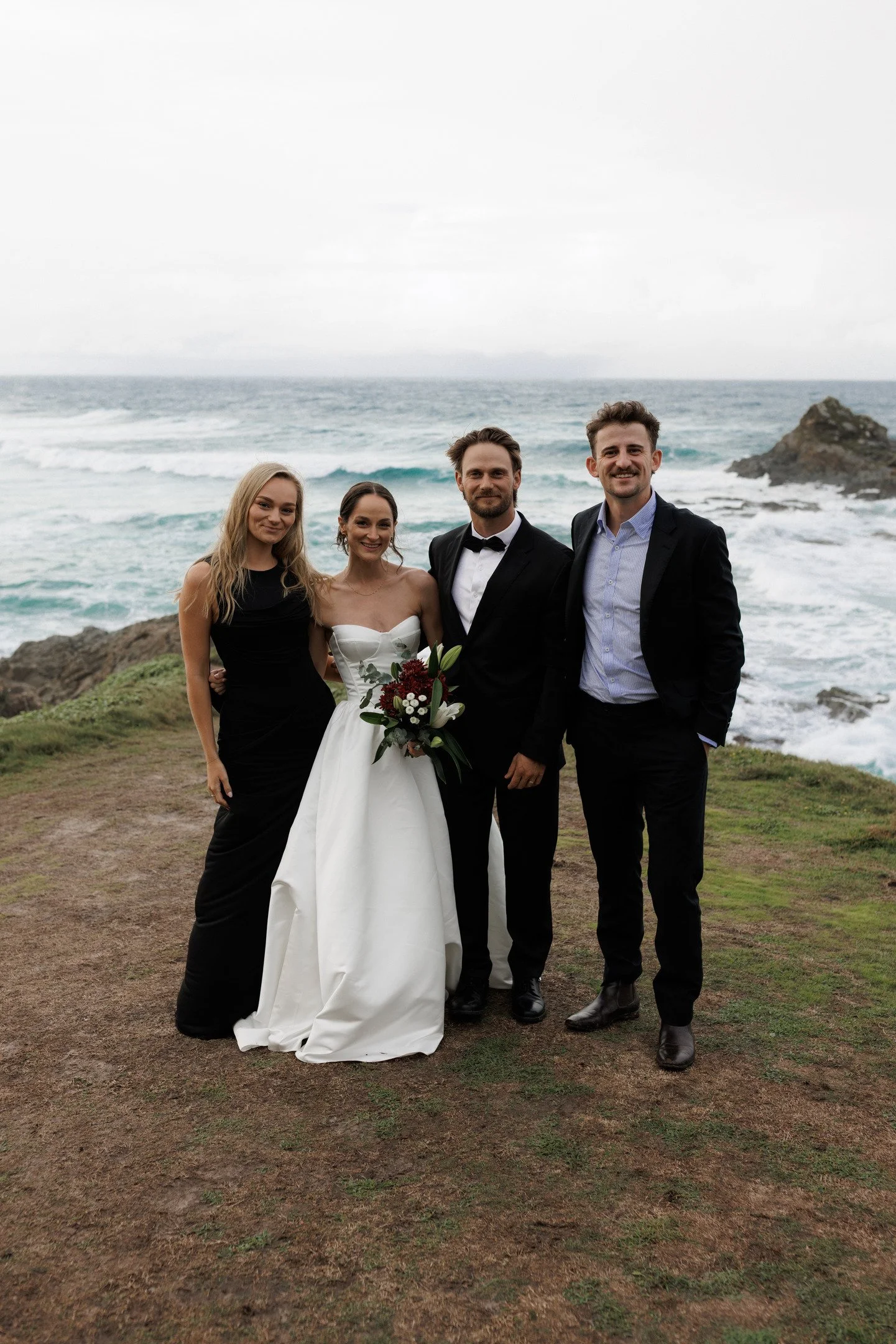 Group of four people standing outdoors near the ocean, dressed in formal attire for a wedding, with rocky shoreline and waves in the background.