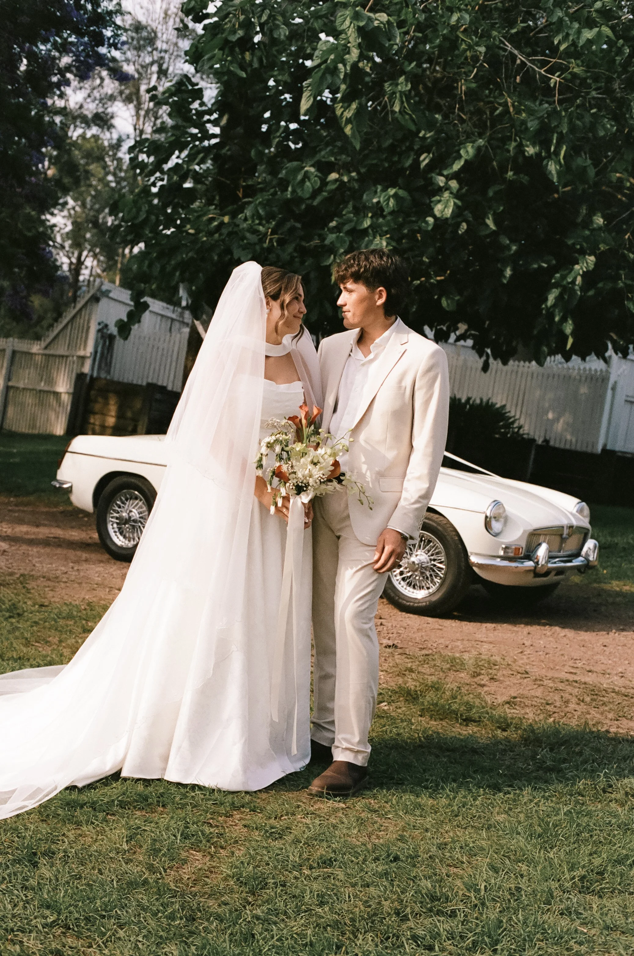 A bride and groom stand close together outdoors under a large tree, holding a bouquet of flowers, with a vintage white convertible car parked behind them.