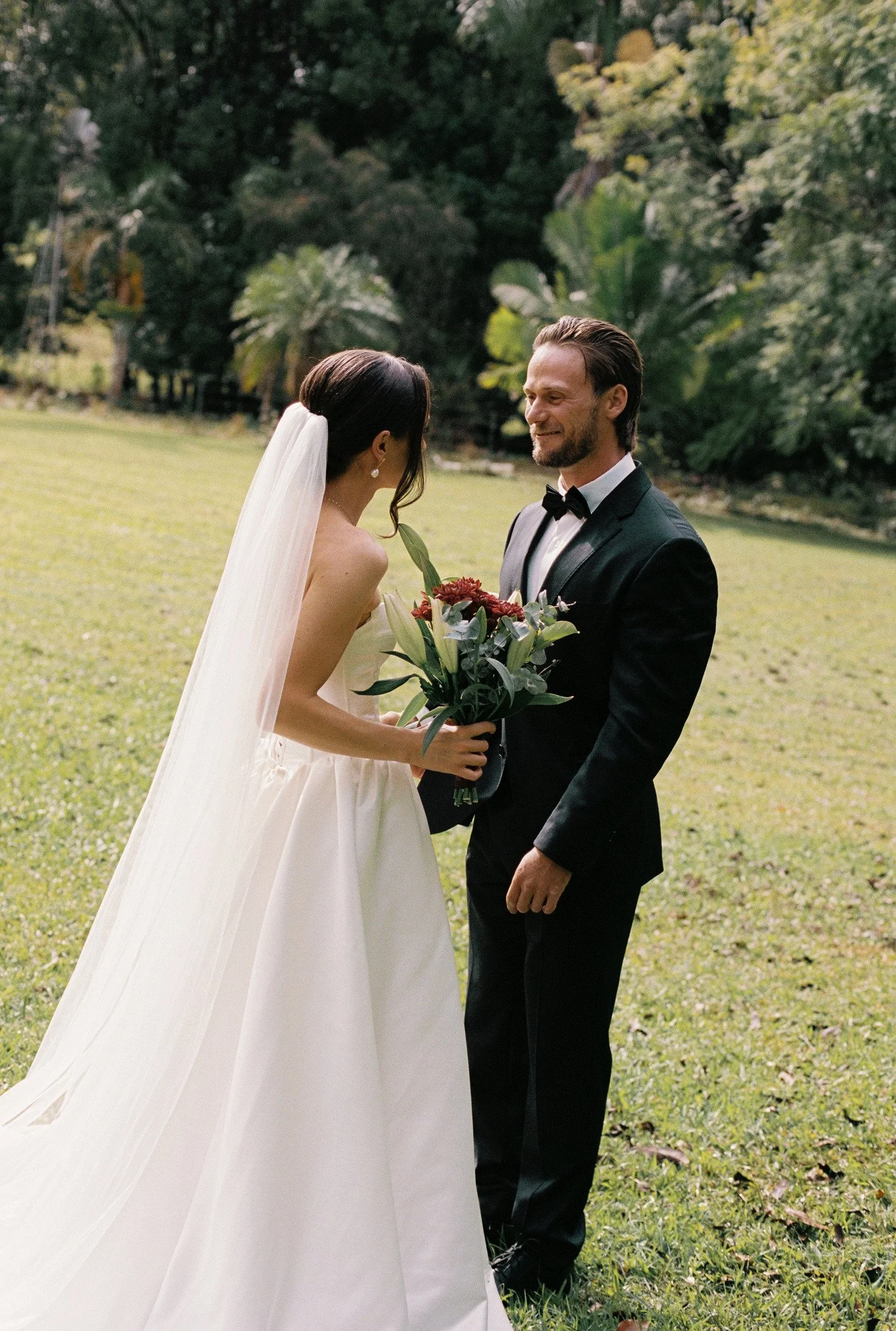 A bride and groom standing on a grassy lawn, facing each other, with the bride holding a bouquet of flowers. The bride is wearing a white wedding dress with a veil, and the groom is in a black tuxedo with a bow tie. They are smiling and looking at ea