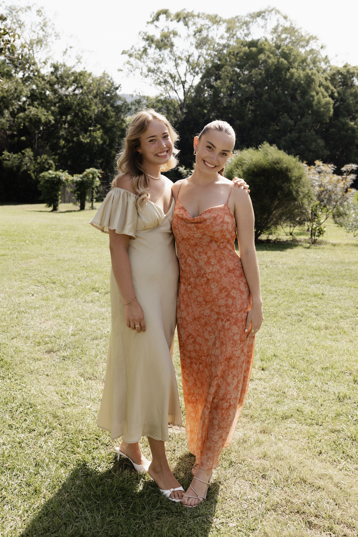 Two young women standing close together outdoors on a sunny day, smiling, with trees and bushes in the background. One is wearing a cream-colored dress with ruffled sleeves, and the other is wearing an orange floral slip dress.