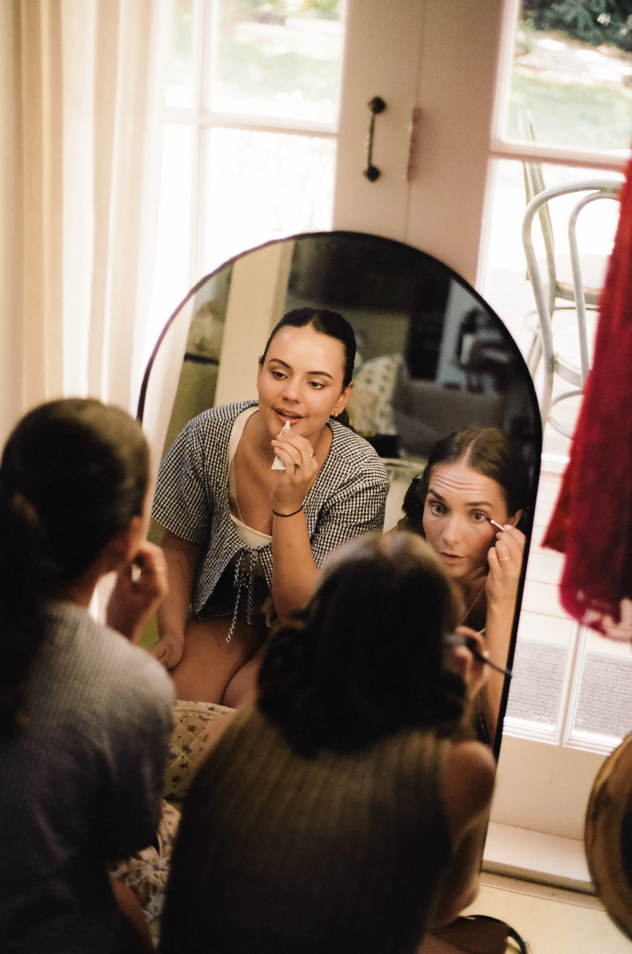 Two women are applying makeup in front of a mirror, with other people nearby, in a cozy room with a large window and natural light.