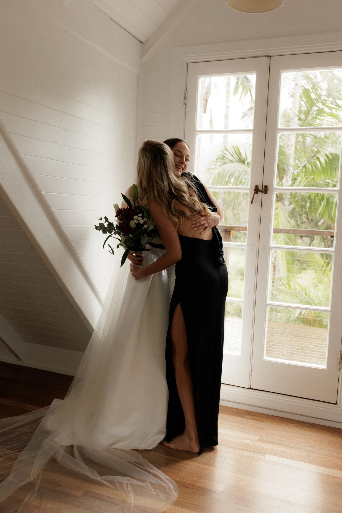 Two women hugging in front of glass doors with greenery outside. One woman is wearing a white wedding dress and holding a bouquet, the other is wearing a black gown with a high slit.