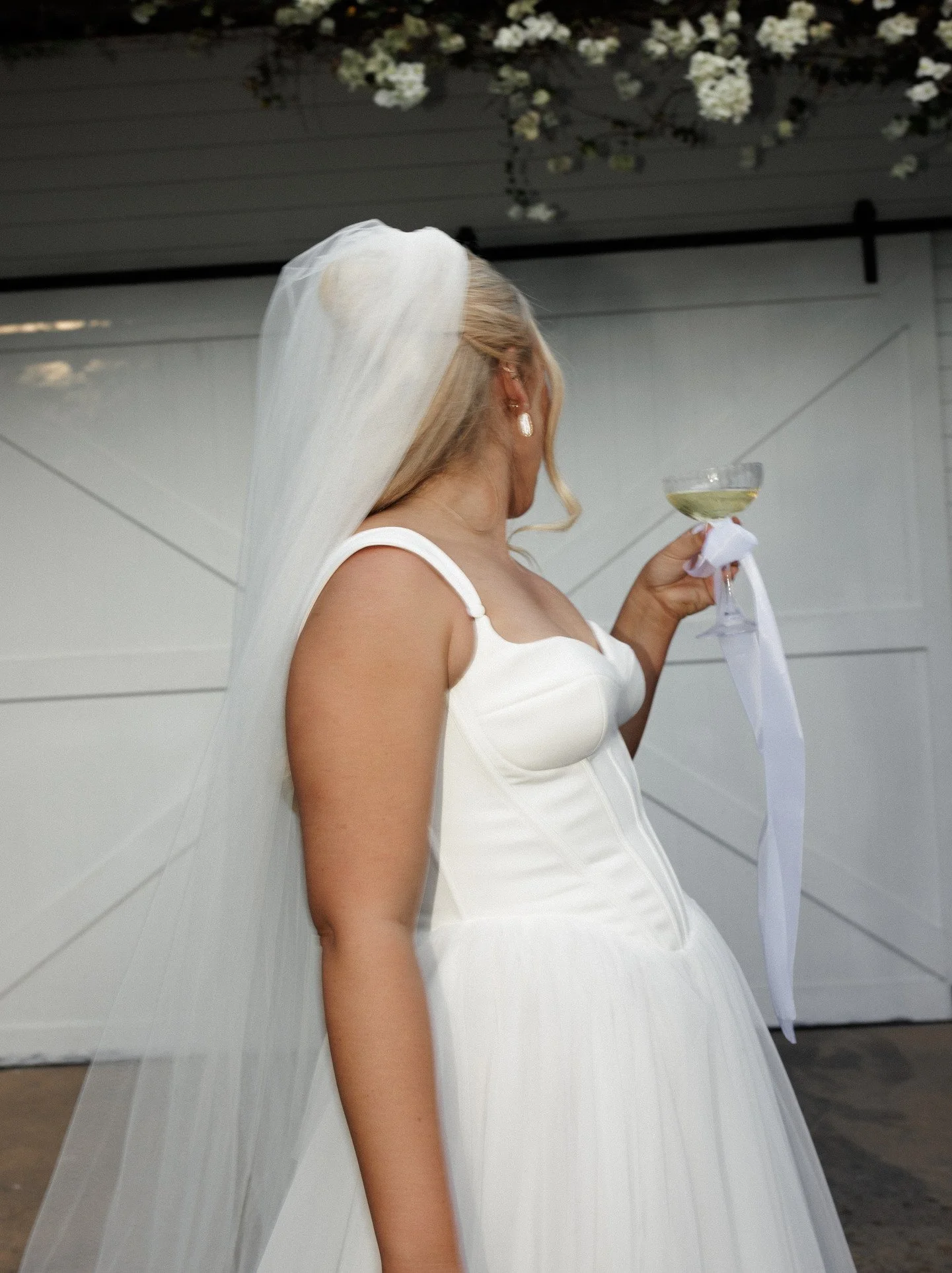 A woman dressed as a bride, wearing a white wedding gown and veil, is holding a glass of champagne and looking away.