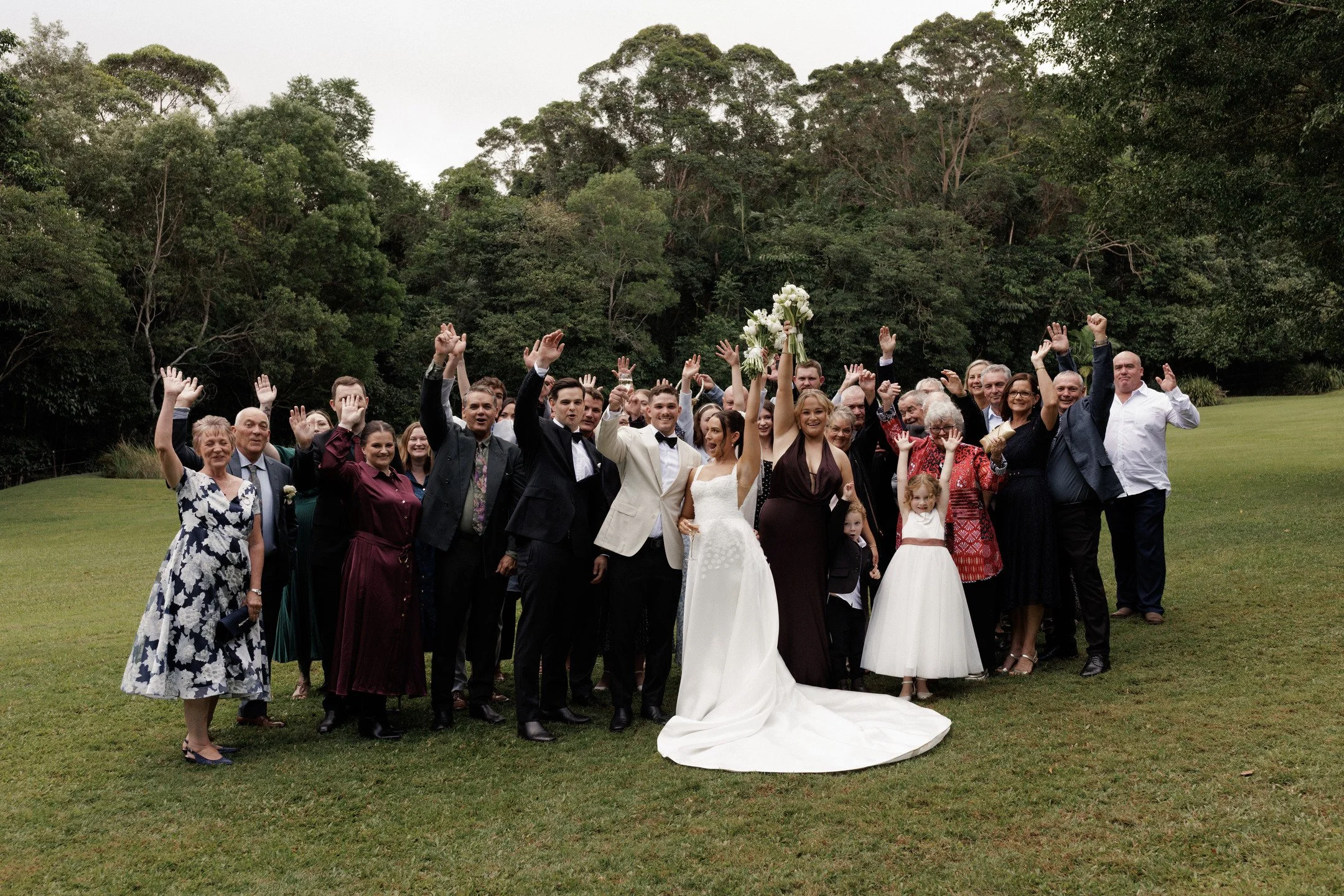 Group of people at a wedding celebration outdoors on a grassy area with trees in the background, some waving and smiling.