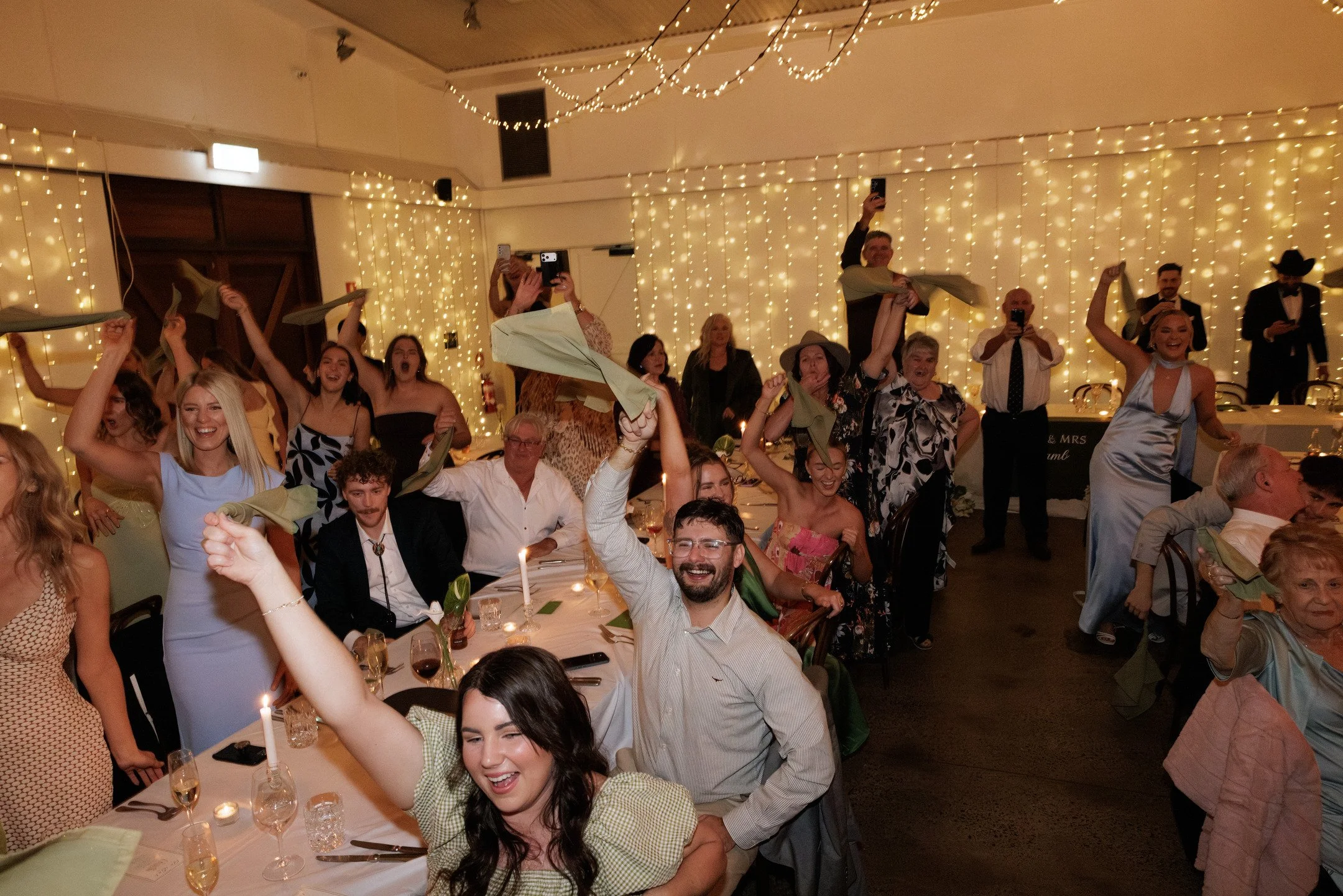 Joyful guests celebrating at a festive event with string lights and elegant attire, some holding green napkins and raising their hands in excitement.