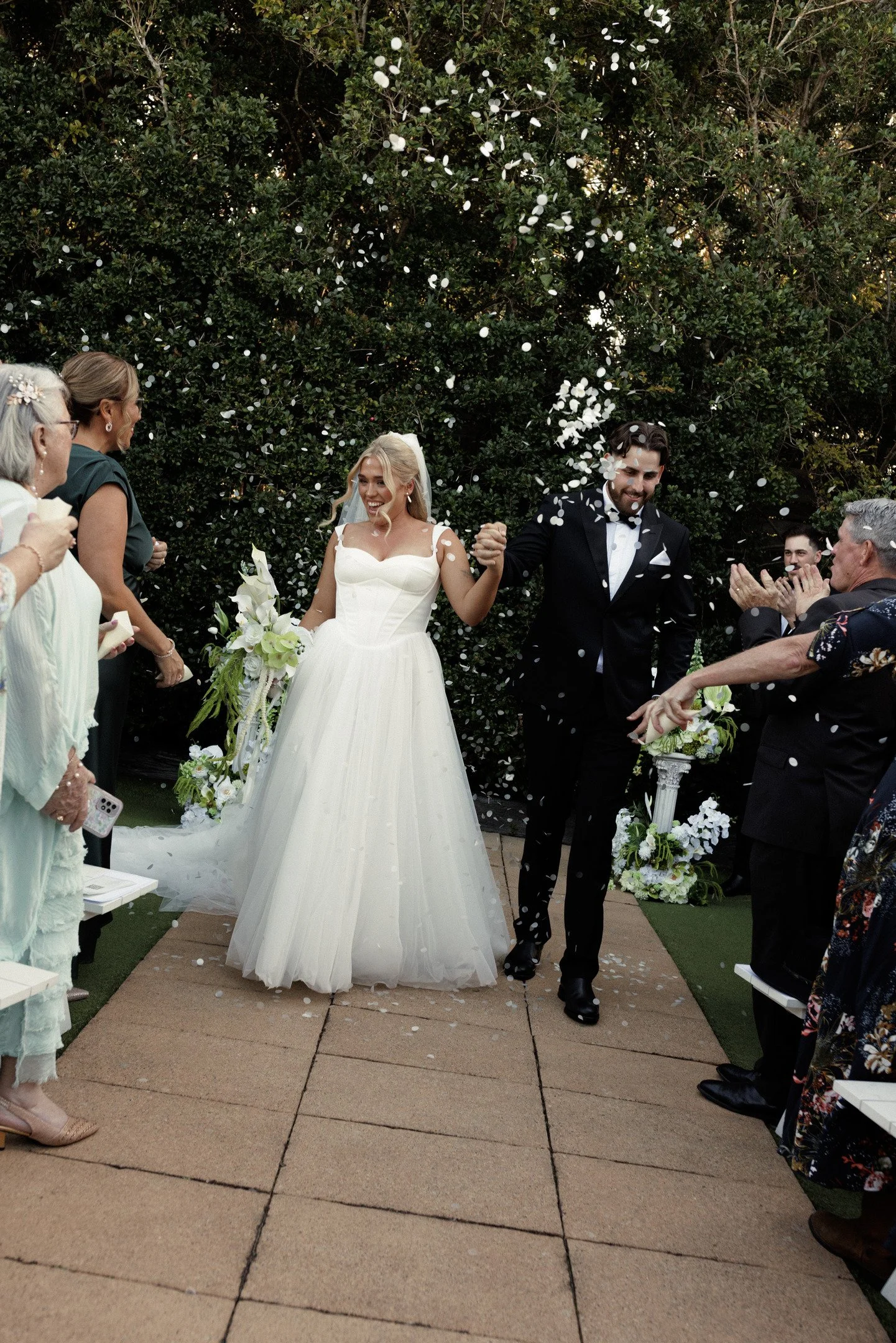Bride and groom holding hands and smiling as they walk down an outdoor aisle, surrounded by wedding guests, with white flower petals falling from above.