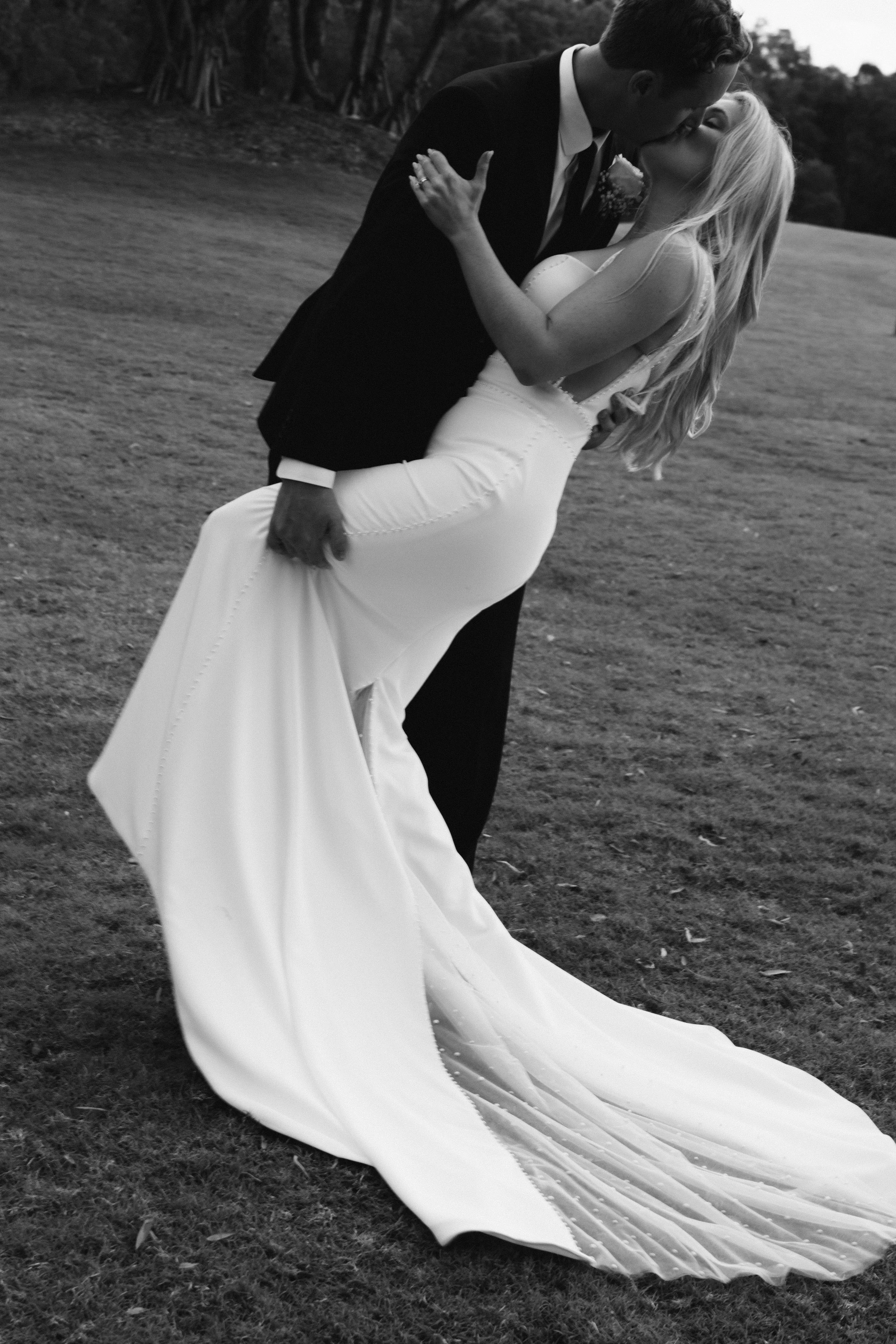 A couple dressed in wedding attire sharing a kiss outdoors, with the groom dipping the bride.