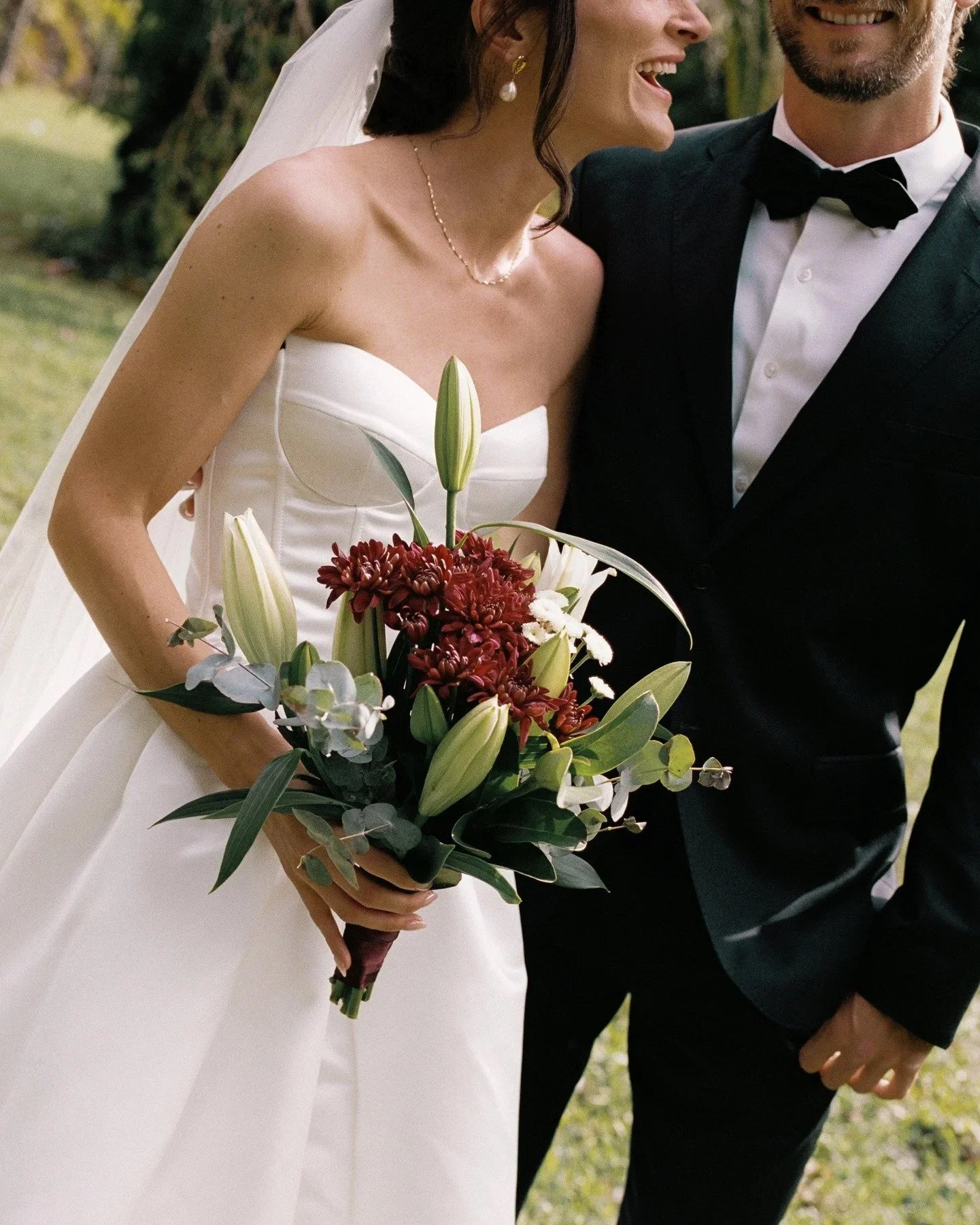 Close-up of a bride in a white wedding gown holding a bouquet of white lilies and red flowers, smiling and talking to a groom in a black tuxedo with a bowtie, outdoors on a sunny day.