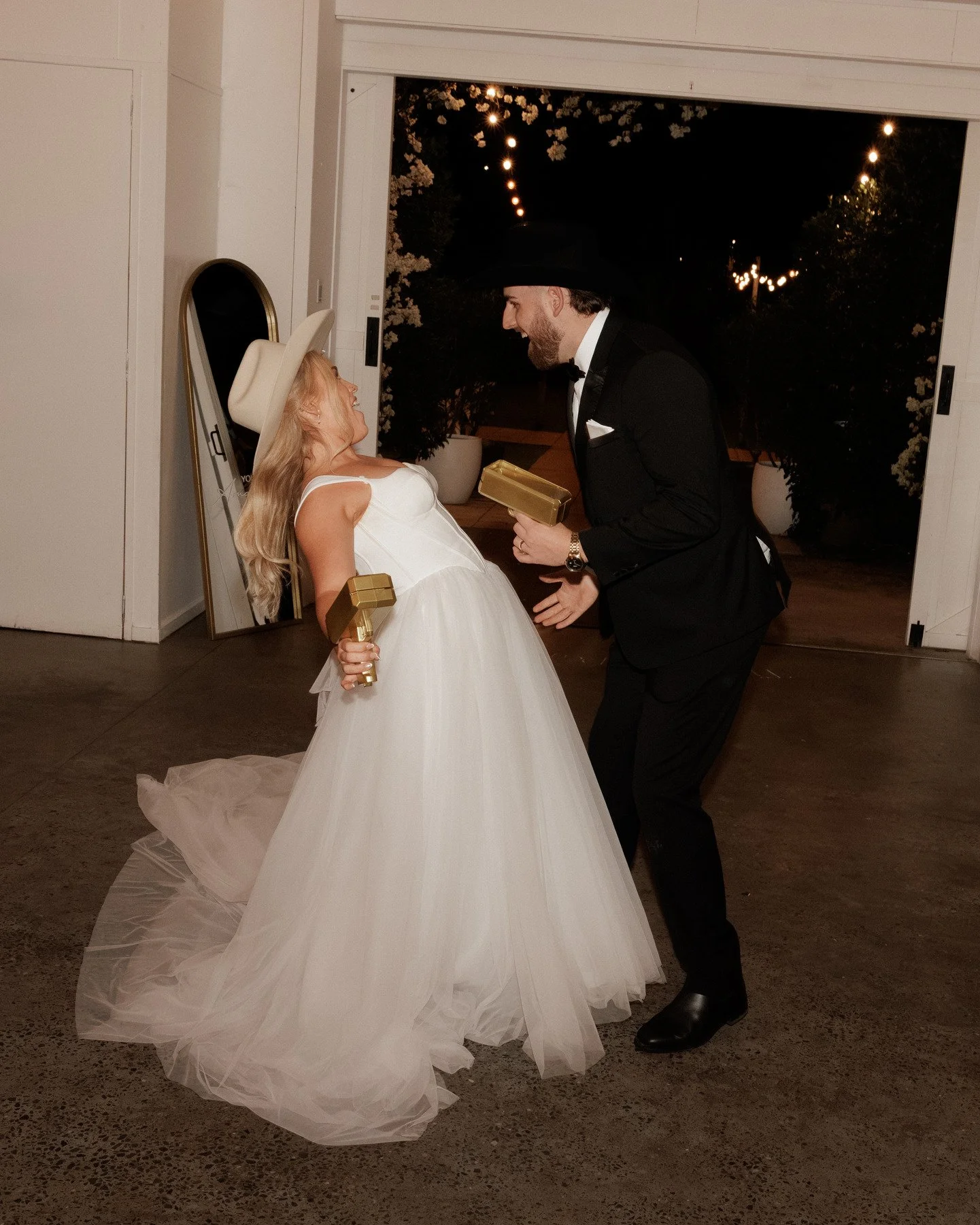 A couple celebrating at a wedding reception, the woman in a white dress and cowboy hat holding a gold gift, the man in a black tuxedo and black cowboy hat smiling at her, both holding gold wrapped gifts, indoors at night.
