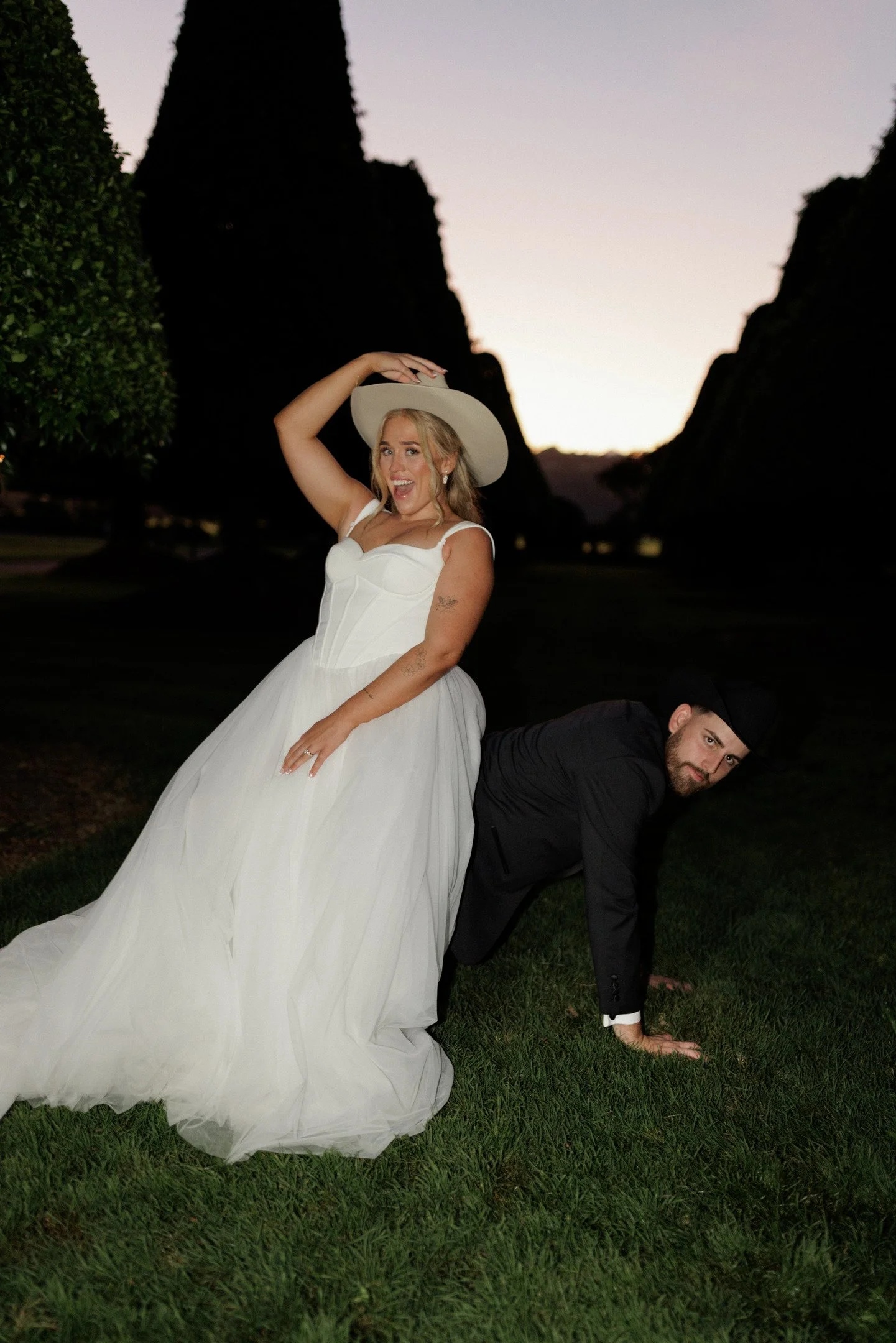 A woman in a white wedding dress and hat is smiling and holding her hat with one hand while leaning on a man in a black suit and hat, who is on all fours on the grass. The background features large dark trees and a sunset sky.