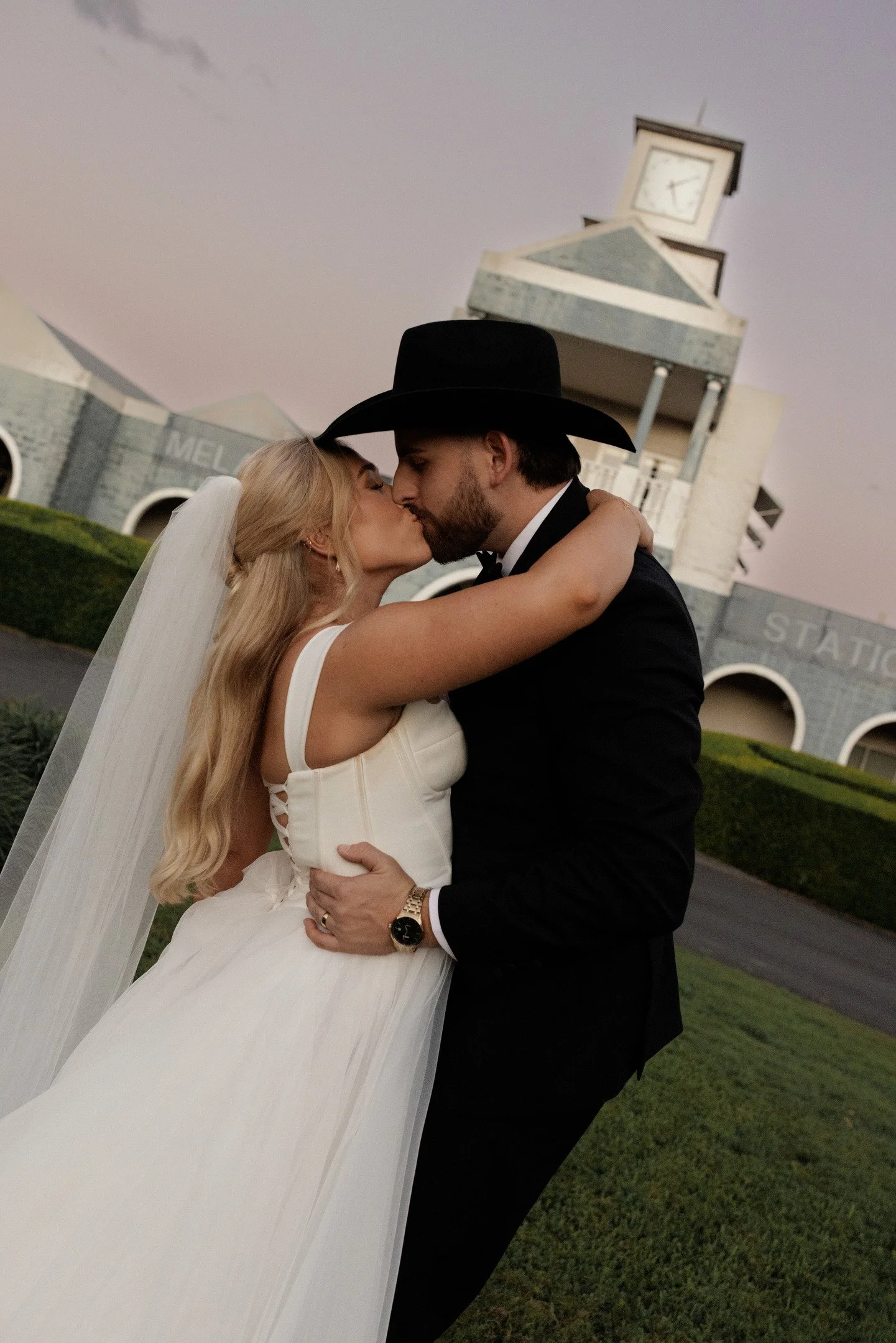 A couple, dressed in wedding attire, kissing outdoors in front of a historic train station with a clock tower, during sunset.