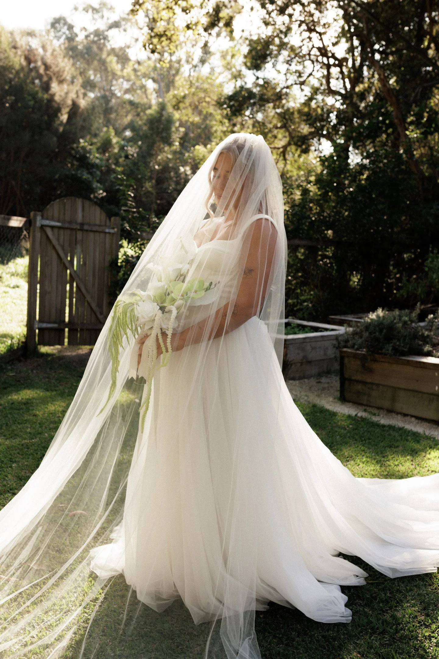 A woman in a wedding dress under a sheer veil stands outside in a garden. She holds a bouquet of white flowers and is surrounded by trees and sunlight.