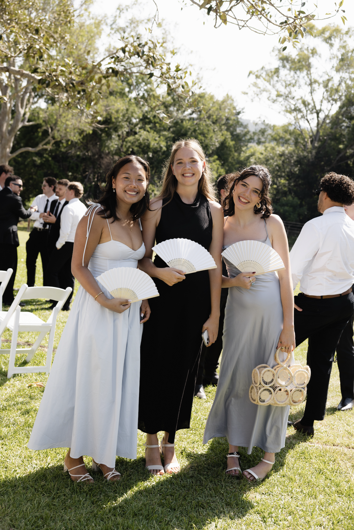 Group of women smiling at an outdoor event, holding fans and bags, dressed in summer attire, with other guests in the background.