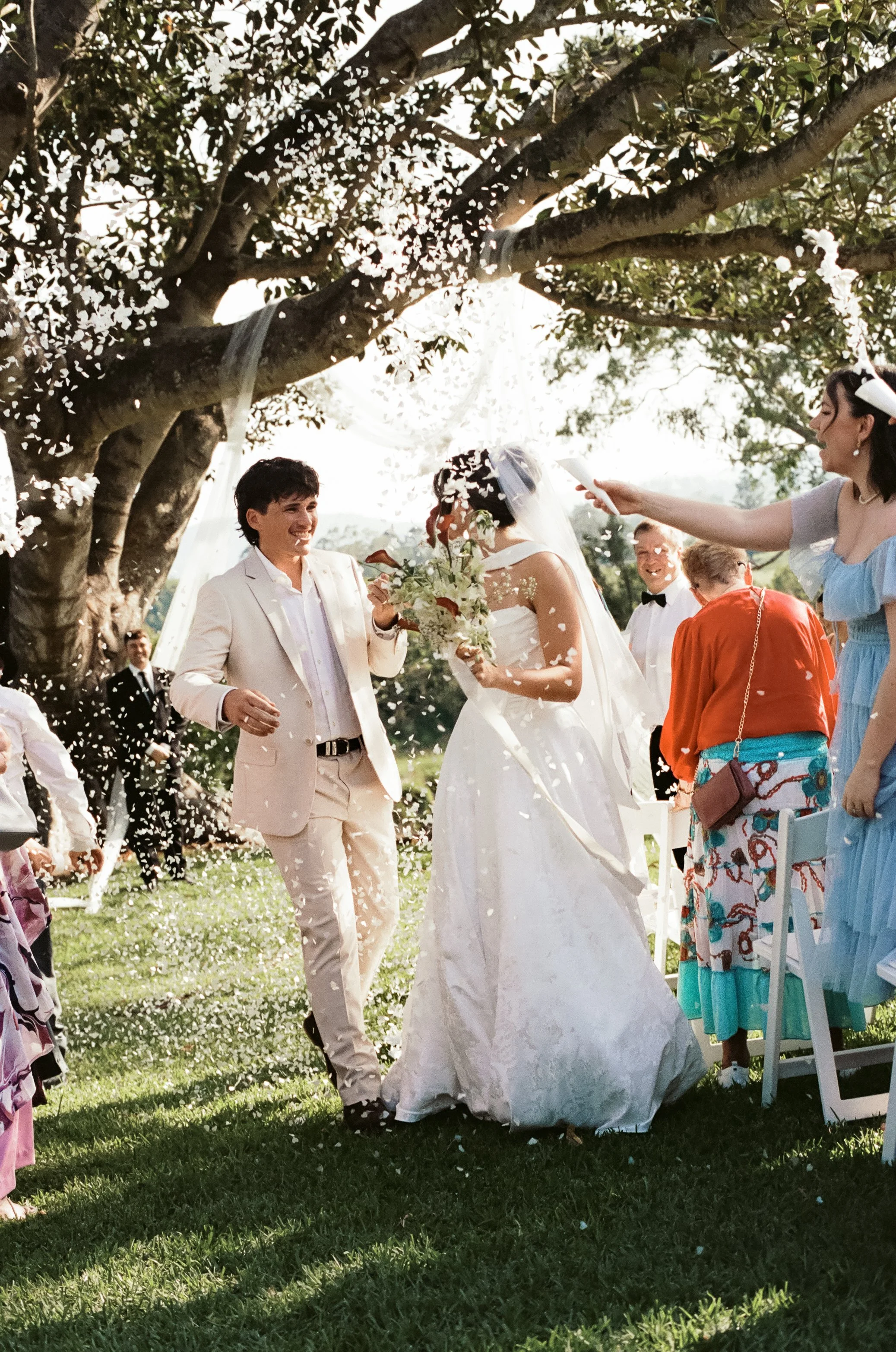 Bride and groom are celebrating outdoors under a large tree, throwing flower petals, with guests around them.