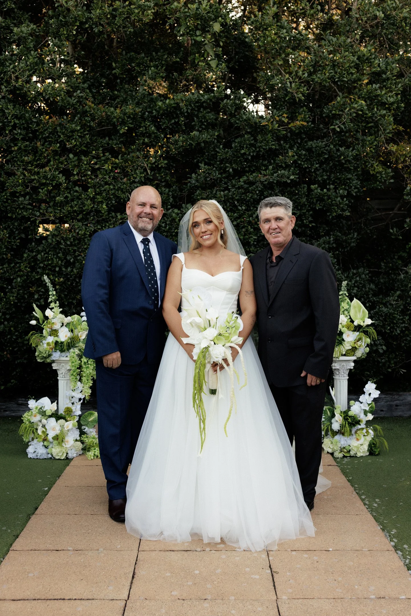 A bride in a white wedding dress holding a bouquet, flanked by two men in dark suits, standing outdoors with a green hedge and floral arrangements behind them.