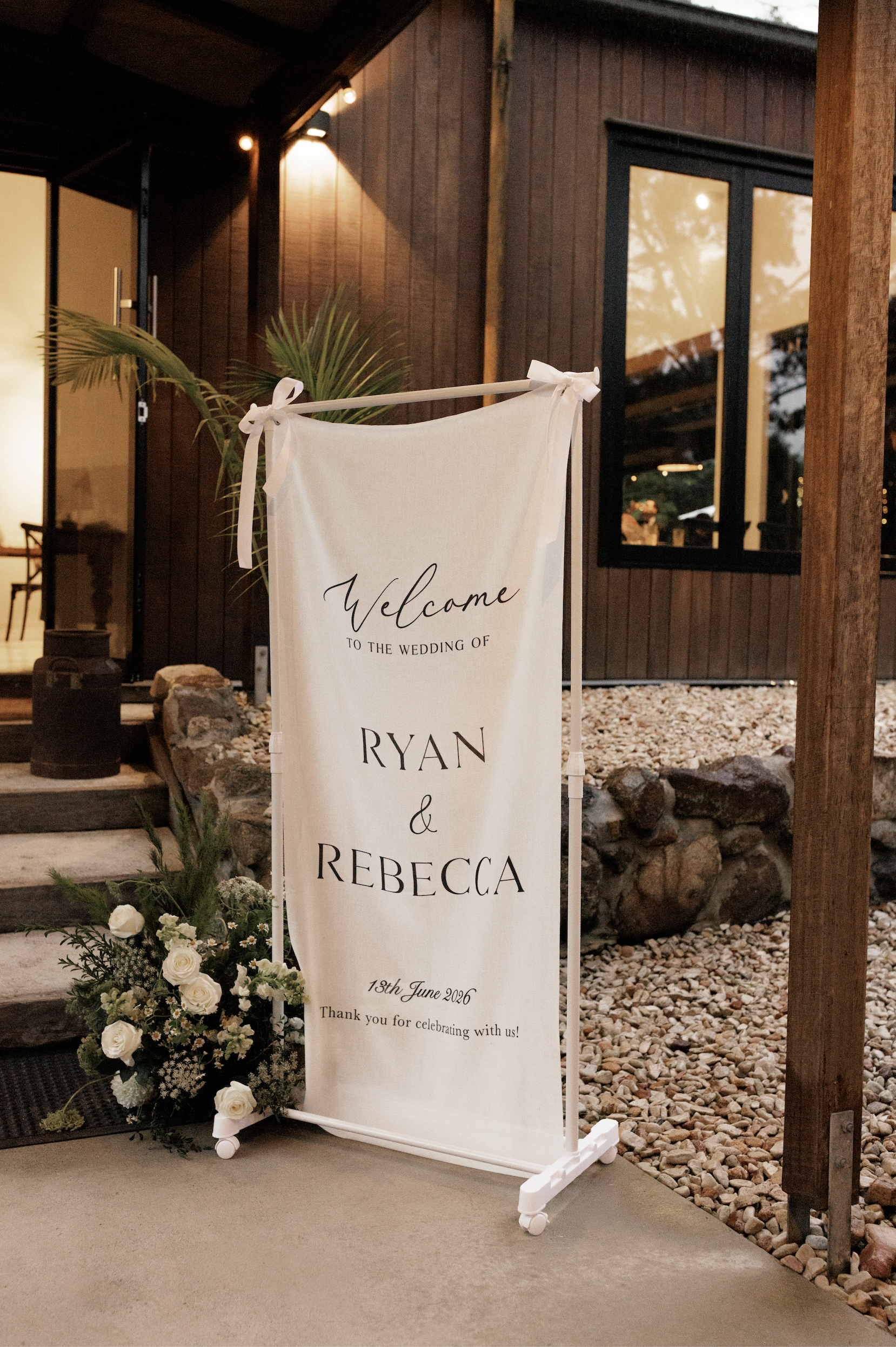 Wedding welcome sign with floral decoration on the ground, outside of a building with wooden walls and glass windows, announcing the wedding of Ryan and Rebecca on June 13, 2026.