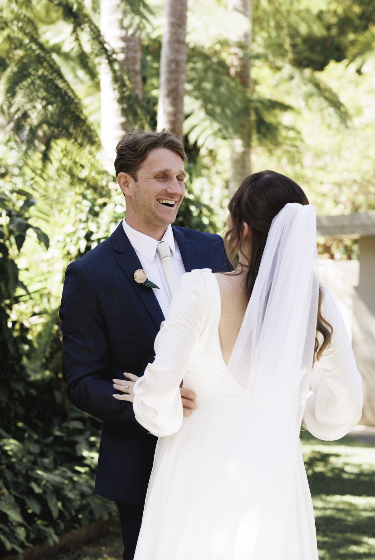 A groom in a navy suit smiling at a bride in a white dress with long sleeves, standing outdoors among greenery and trees, during their wedding ceremony.