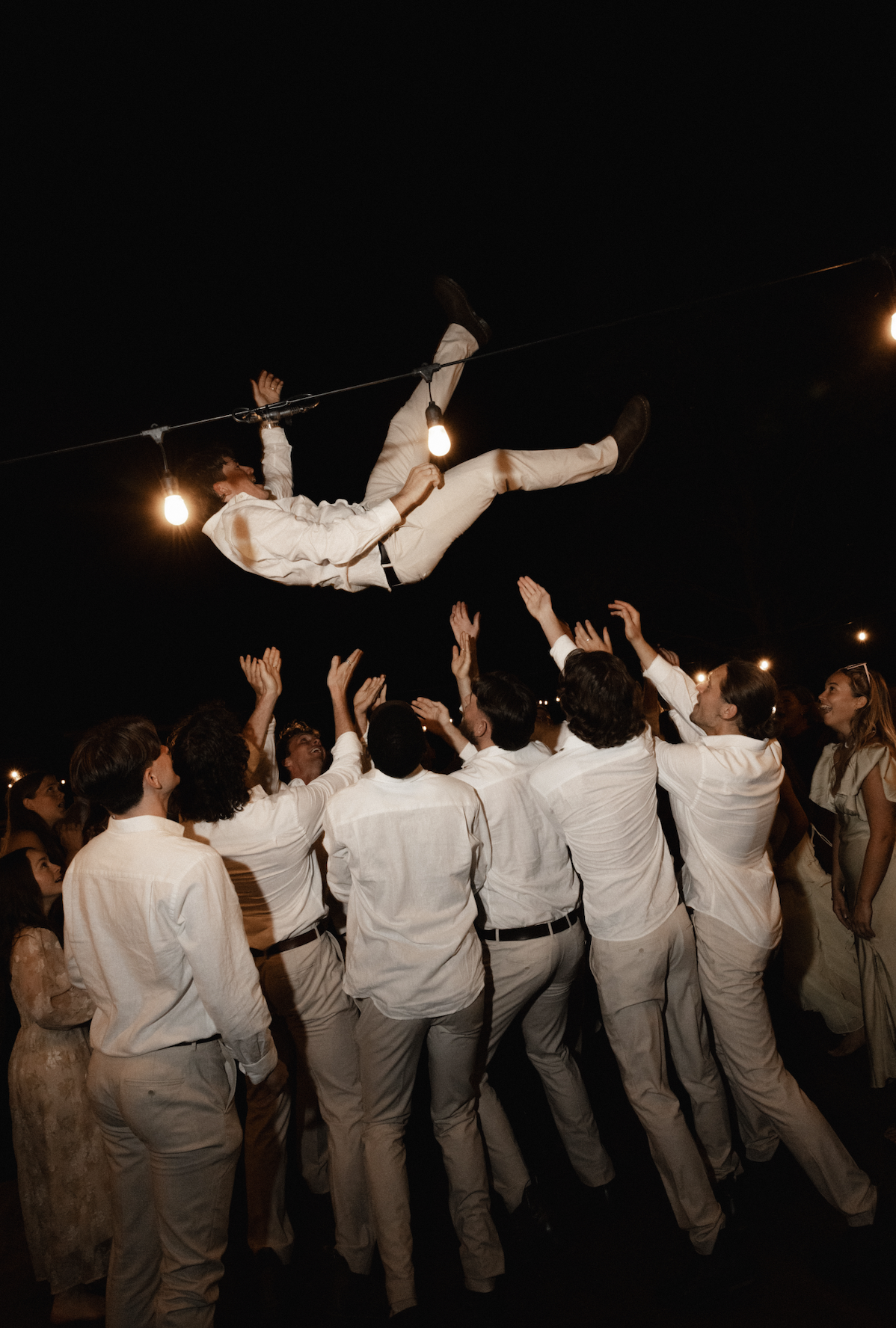 Group of people at night, participating in a traditional dance, tossing a man in a white outfit into the air under string lights.