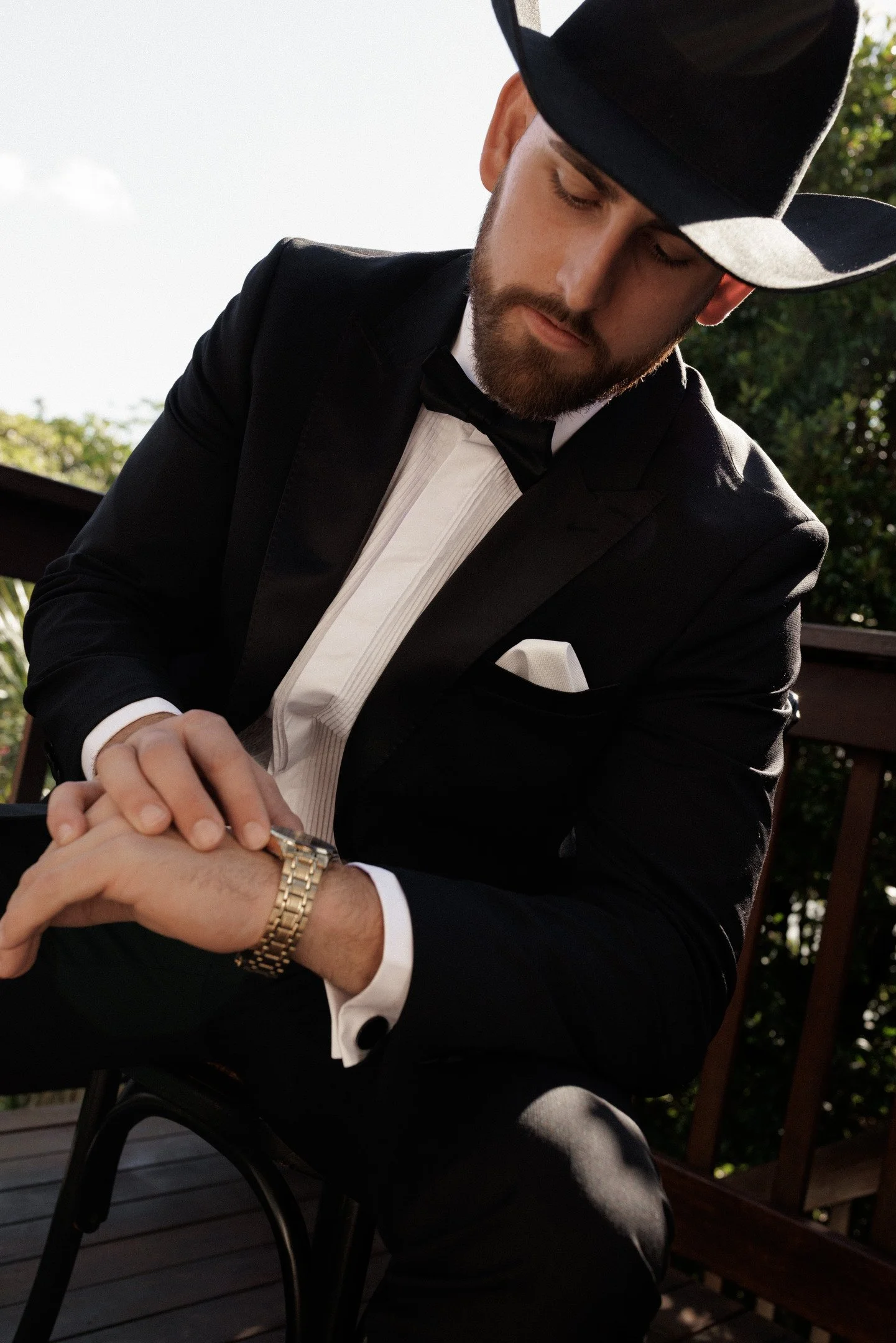 Man in black tuxedo and bow tie wearing a black wide-brimmed hat, looking down, sitting on a bench outdoors with green foliage in the background.