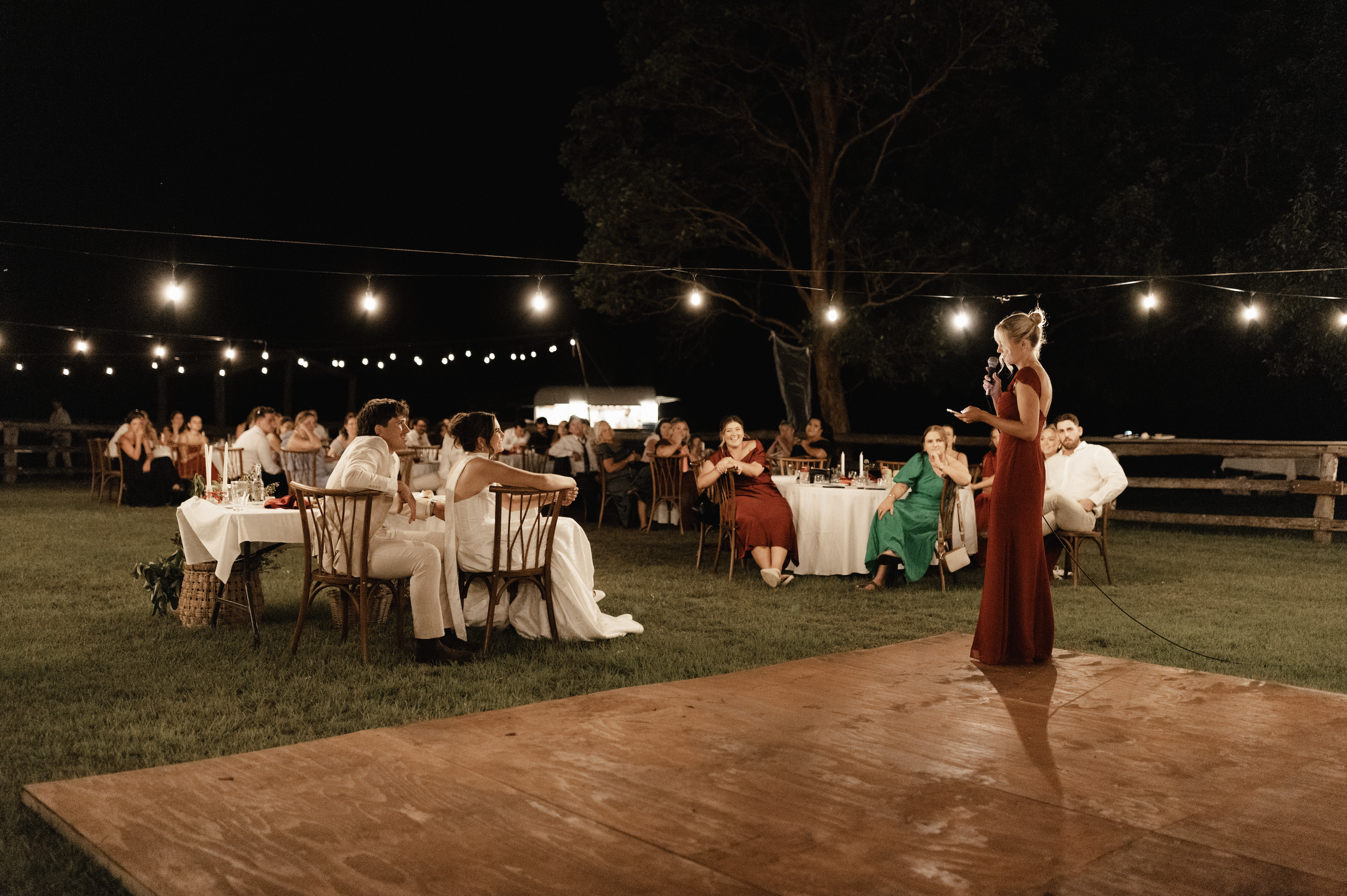 Nighttime outdoor wedding reception with guests sitting at tables under string lights, a woman in a red dress giving a speech on a small wooden dance floor.