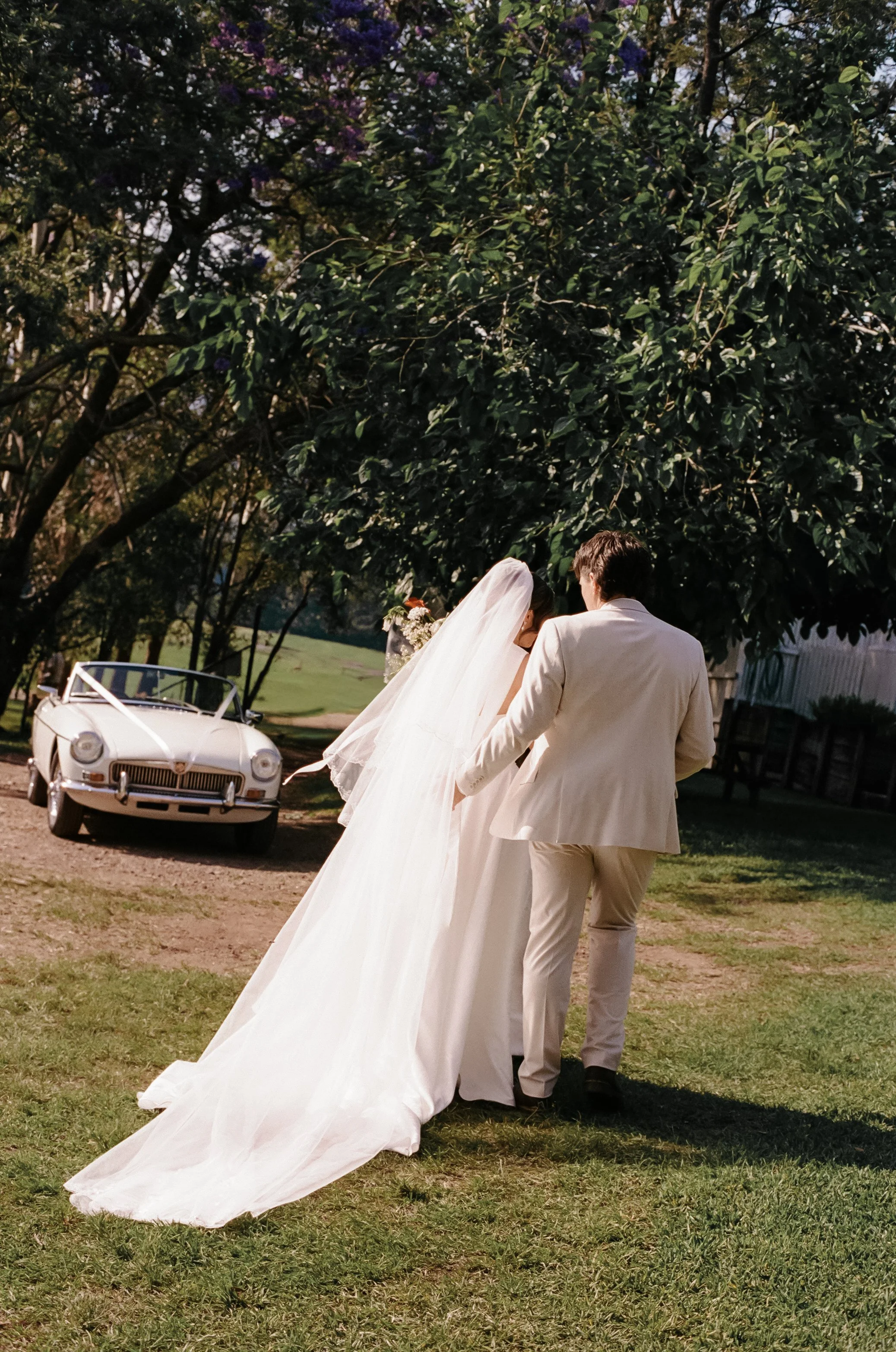 A bride and groom walking together outdoors, with a vintage white car decorated with ribbon in the background, trees, and grassy area.