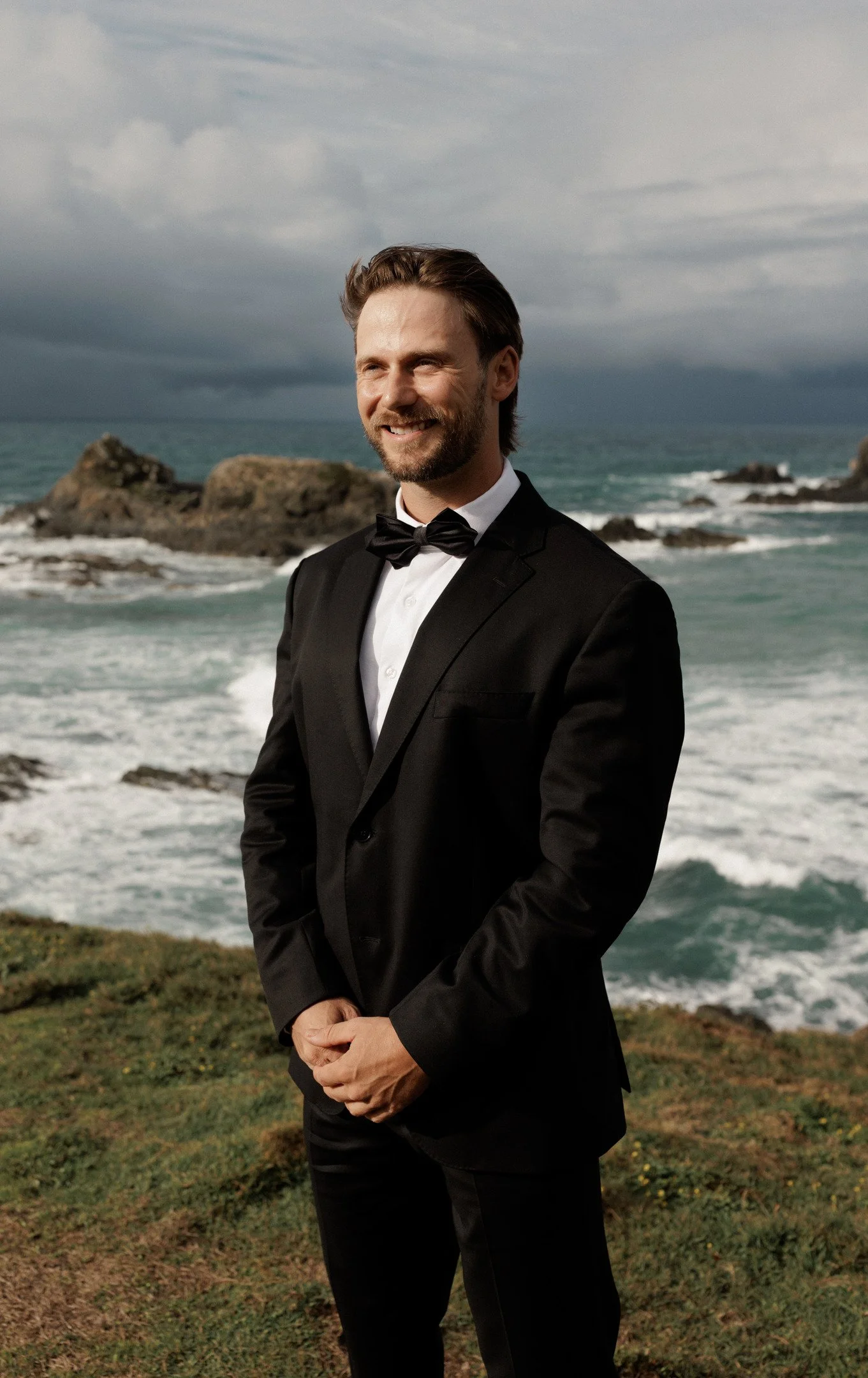 A man in a tuxedo standing outdoors near the ocean, with rocky formations and stormy clouds in the background.