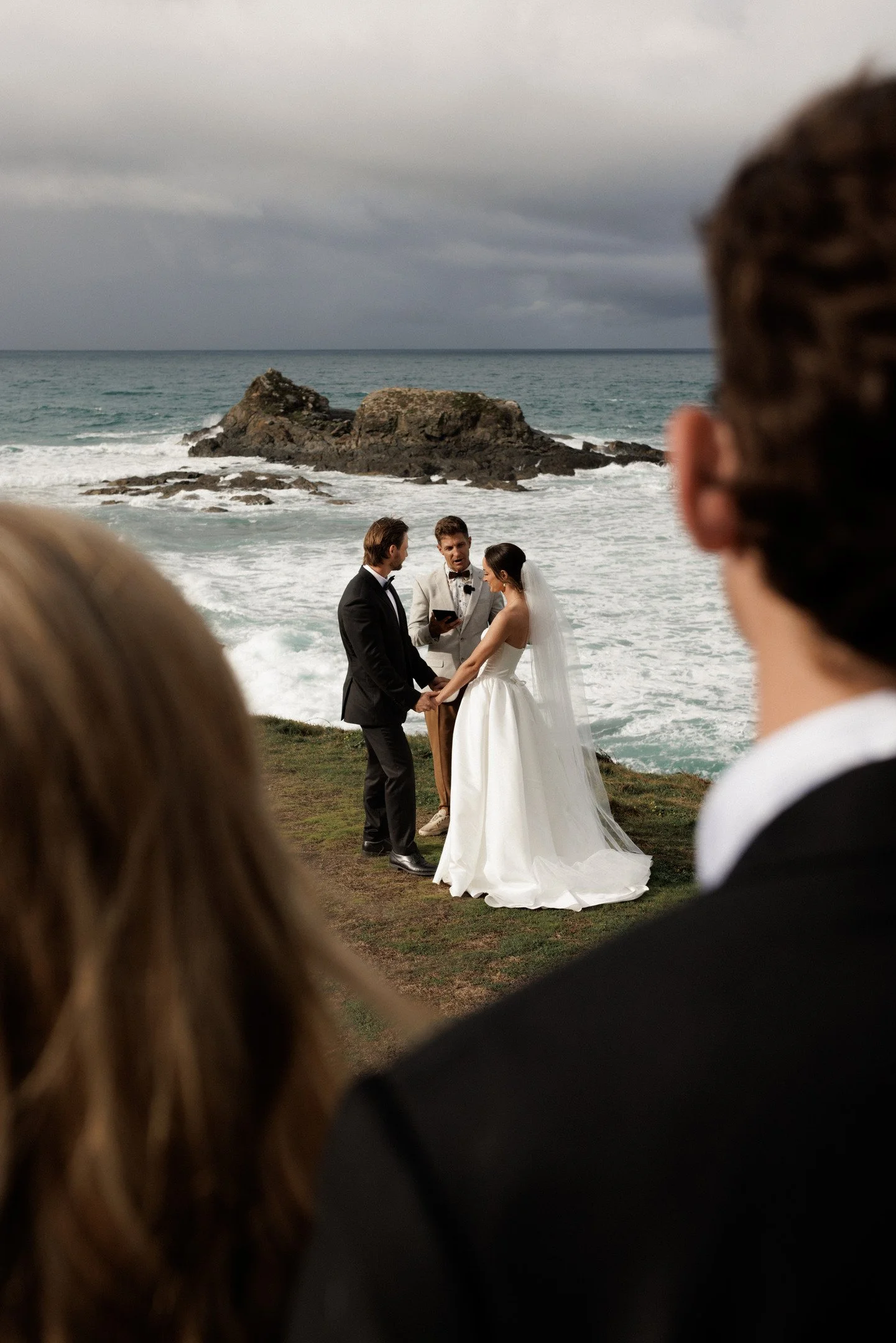 A wedding ceremony on the beach with the bride and groom holding hands, an officiant reading vows, and the ocean with rocks in the background.