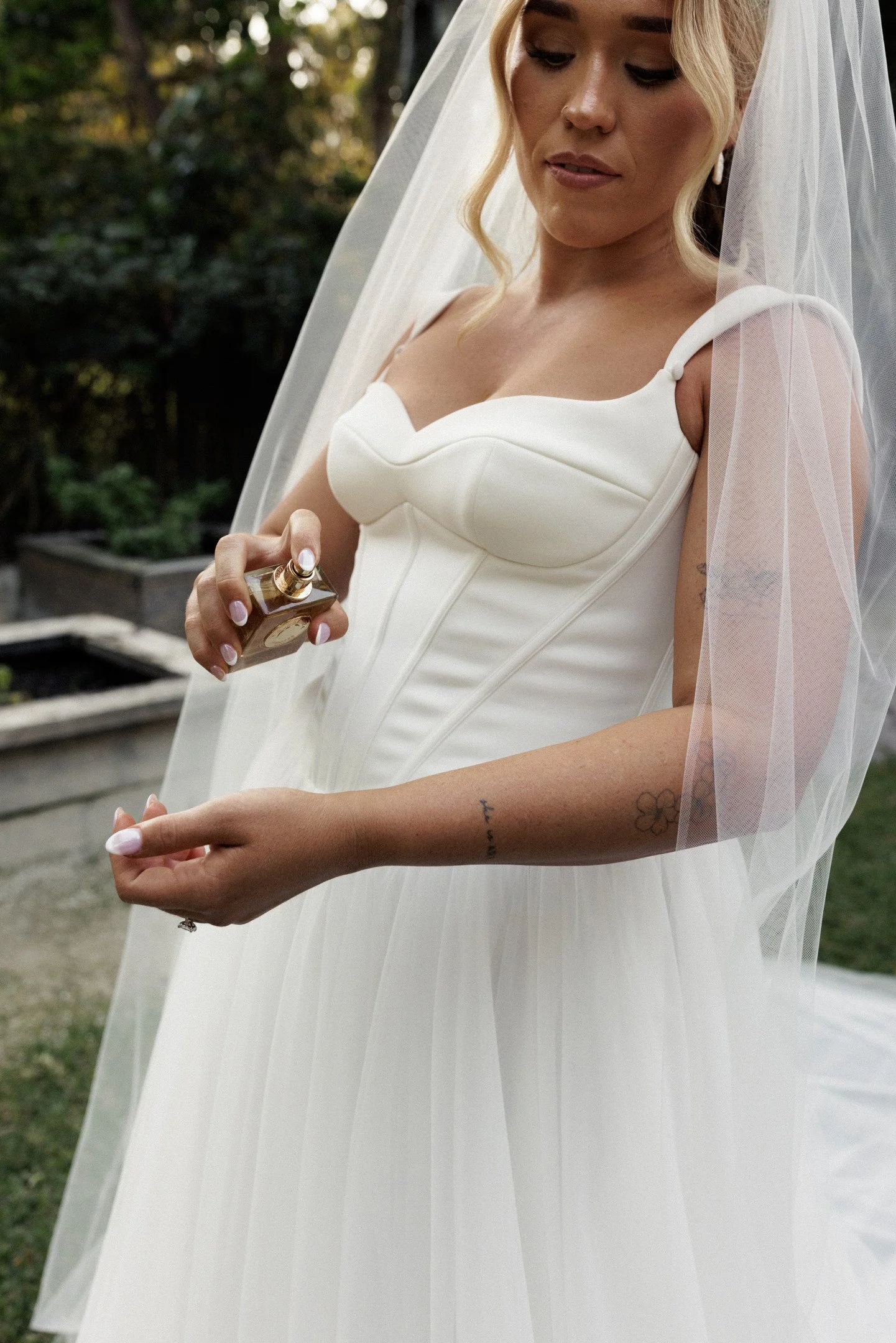 A bride in a white wedding dress with a veil holding a perfume bottle outdoors.
